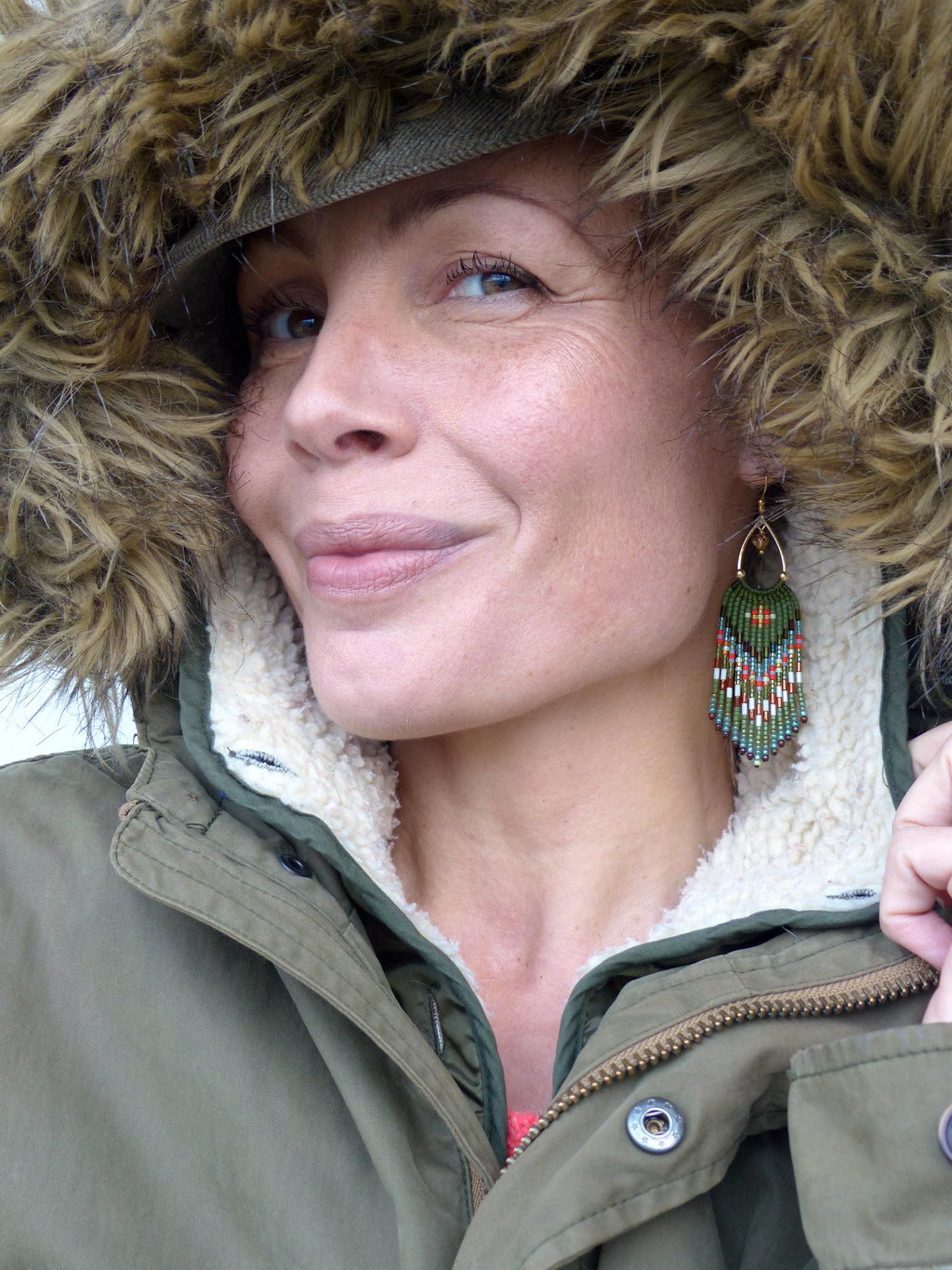 Peut inclure: Une femme porte une parka verte avec une capuche doublée de fourrure et des boucles d'oreilles en perles. Les boucles d'oreilles sont vertes, rouges et blanches, avec un crochet doré. La parka a une doublure blanche et moelleuse.