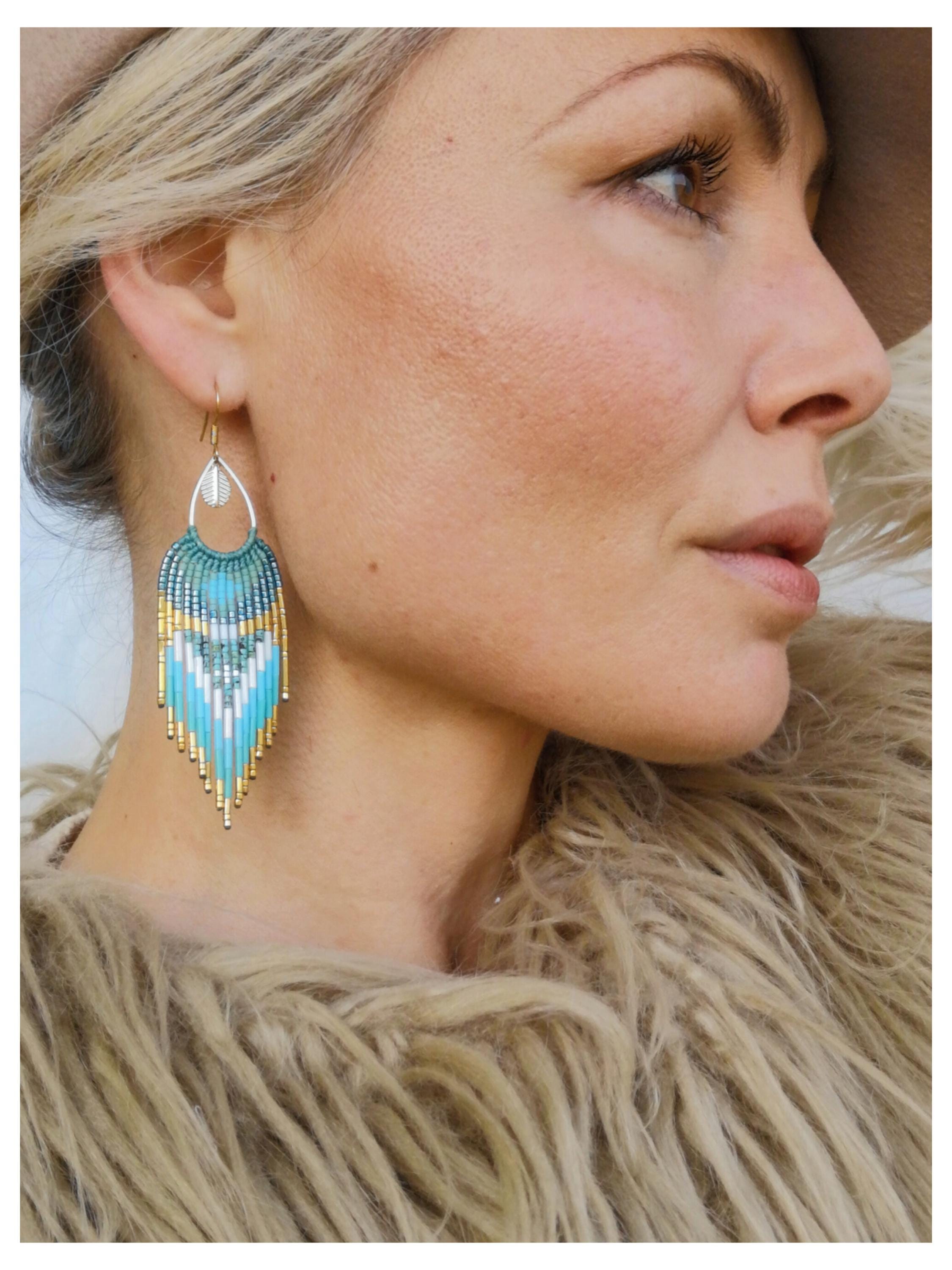 Peut inclure: Boucle d'oreille perlée avec une partie supérieure argentée en forme de goutte d'eau et une frange perlée turquoise, blanche et dorée. La boucle d'oreille est suspendue à un crochet doré. La boucle d'oreille est portée par une personne aux cheveux blonds et un manteau de fourrure beige.