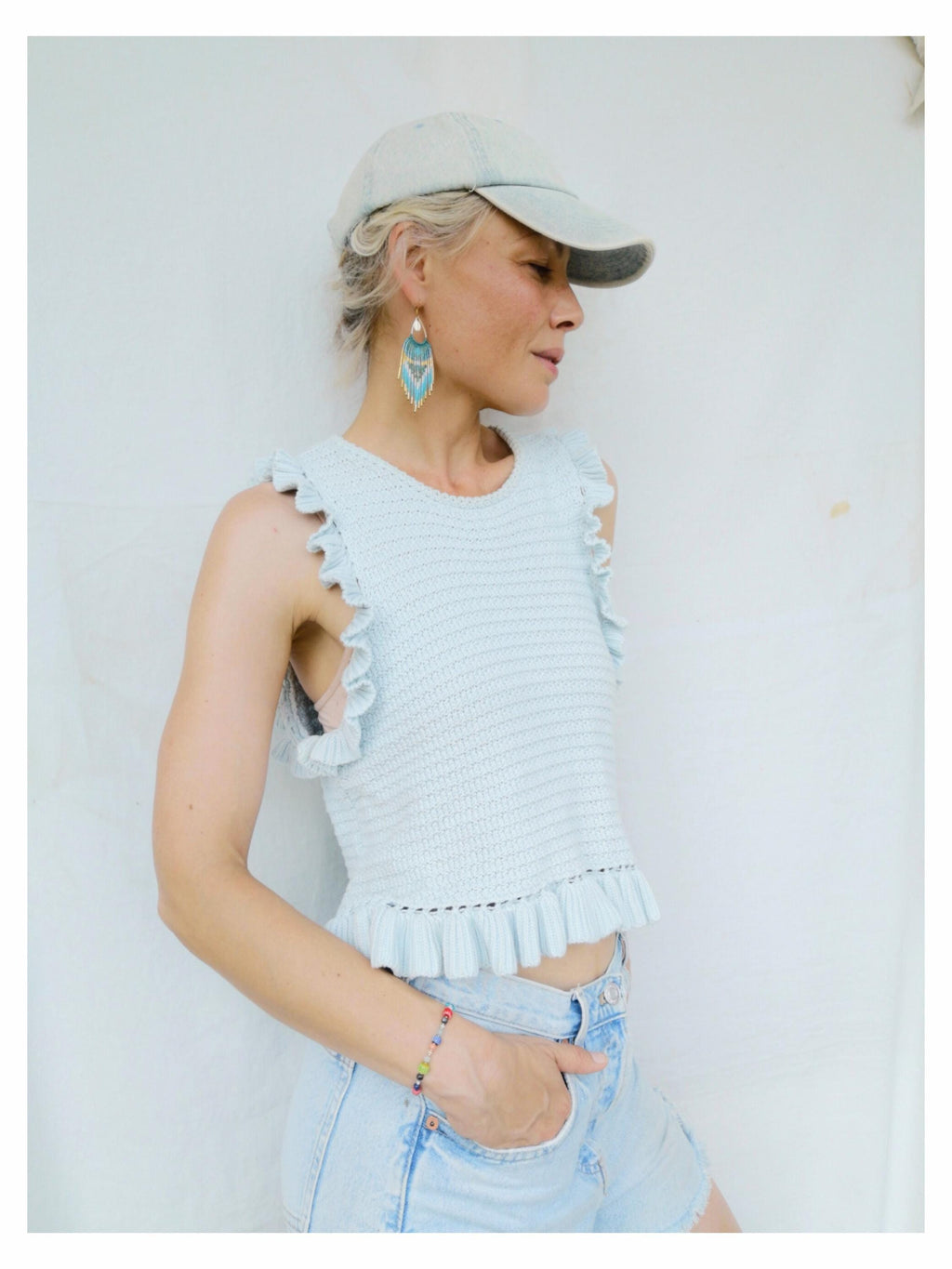 Peut inclure: Haut en crochet bleu clair avec des bords à volants et une casquette de baseball assortie. Le modèle porte également un short en jean délavé, un bracelet de perles colorées et des boucles d'oreilles turquoise et dorées. La tenue est décontractée et estivale.