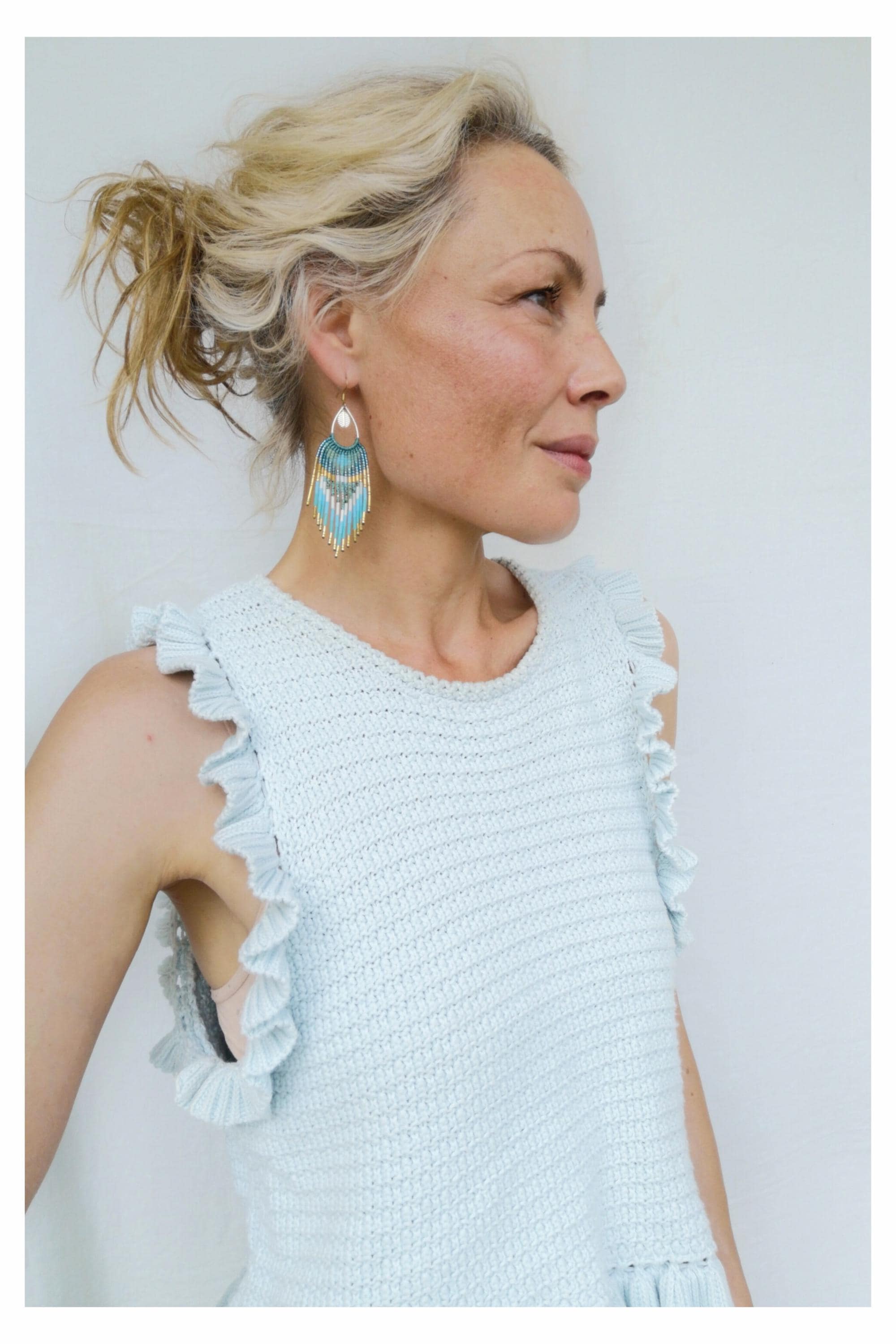 Peut inclure: Une femme porte un haut en tricot bleu clair avec des manches à volants et des boucles d'oreilles en perles. Les boucles d'oreilles ont un dessus doré avec des franges turquoise et dorées. La femme a les cheveux blonds attachés.