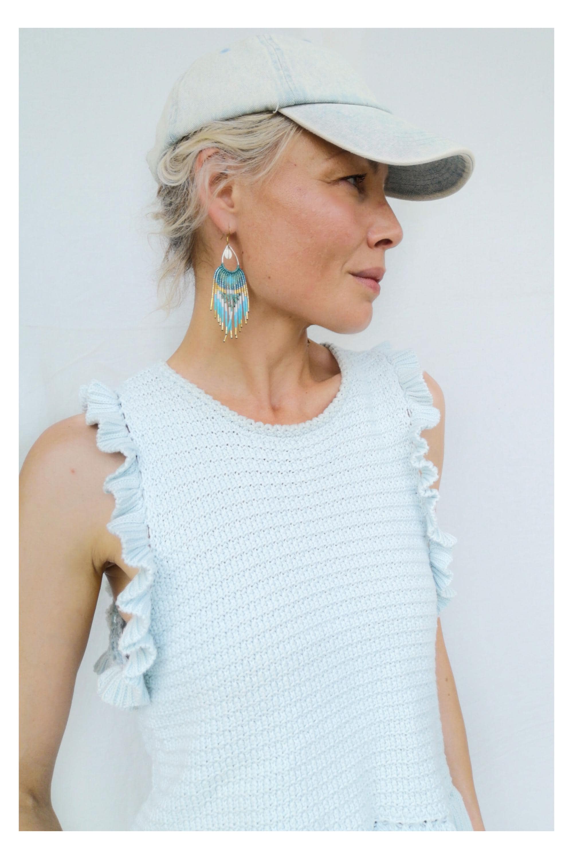 Peut inclure: Haut en tricot bleu clair avec des manches à volants et une casquette de baseball assortie. La femme porte des boucles d'oreilles en perles turquoise et dorées. Les boucles d'oreilles ont une forme de goutte d'eau avec un design à franges.