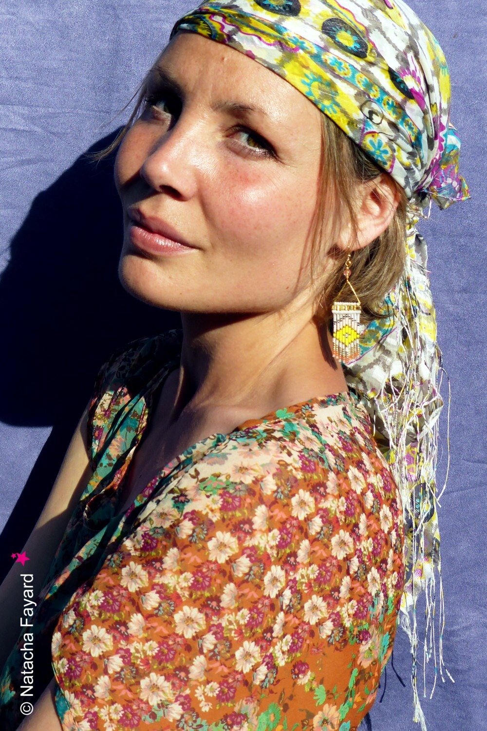 Peut inclure: Une femme aux longs cheveux bruns portant une robe à imprimé floral et un foulard à motifs colorés. Elle porte de longues boucles d'oreilles en perles avec des accents jaunes et blancs.