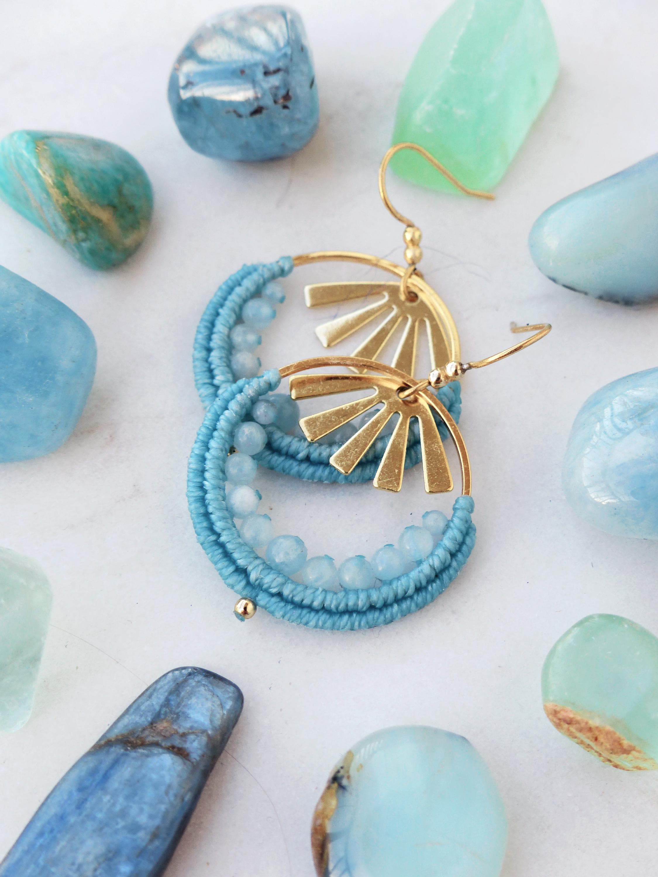 Peut inclure: Boucles d'oreilles dorées en forme de cerceau avec un motif de soleil, ornées de macramé bleu clair et de petites perles bleu clair. Les boucles d'oreilles sont entourées de diverses pierres précieuses bleues et vertes sur une surface blanche.