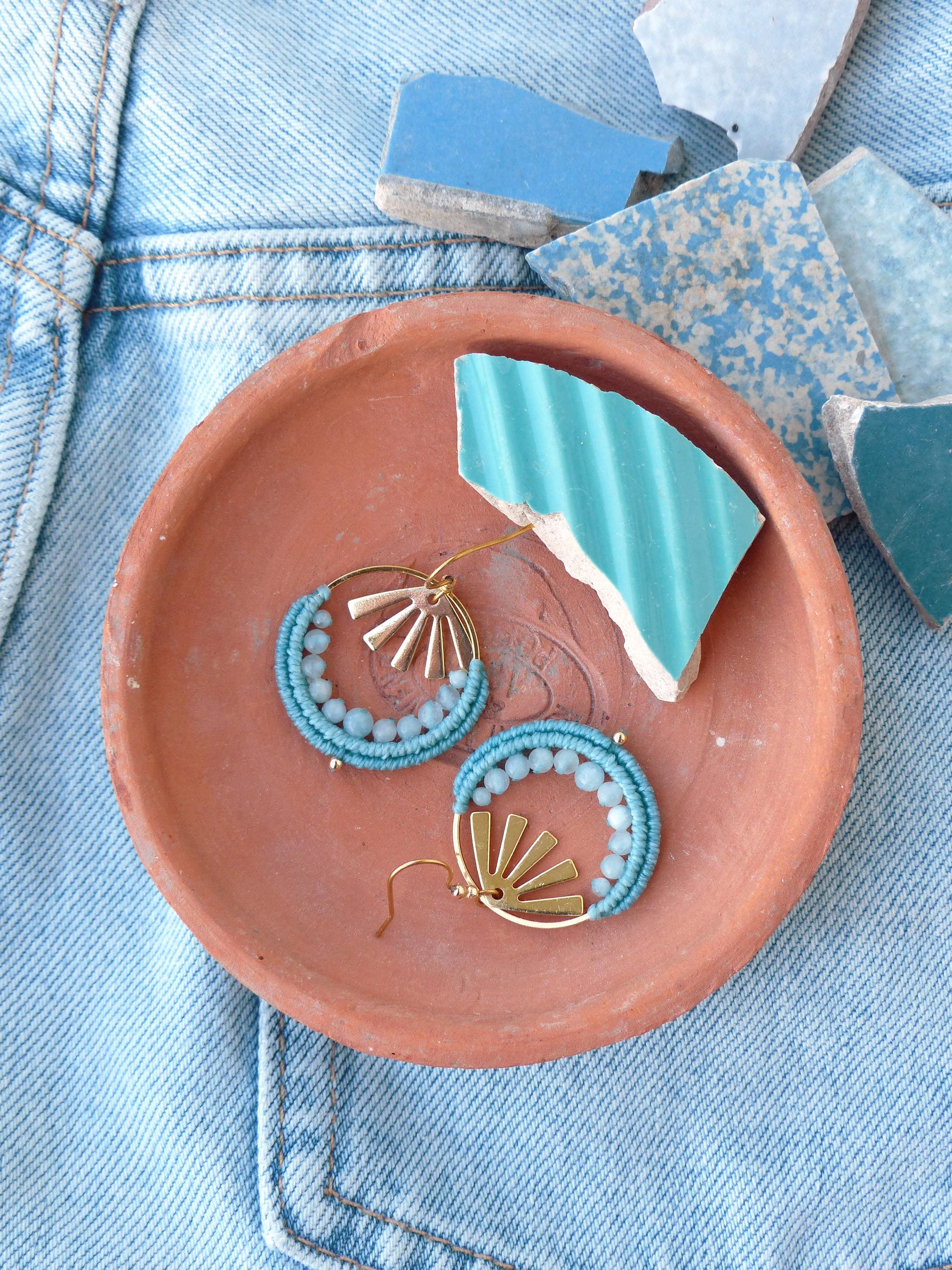 Peut inclure: Boucles d'oreilles faites à la main avec un motif de soleil doré et des perles bleu clair. Les boucles d'oreilles sont présentées sur une petite coupelle en terre cuite, avec des morceaux de carrelage bleu et blanc. Le fond est en tissu denim.