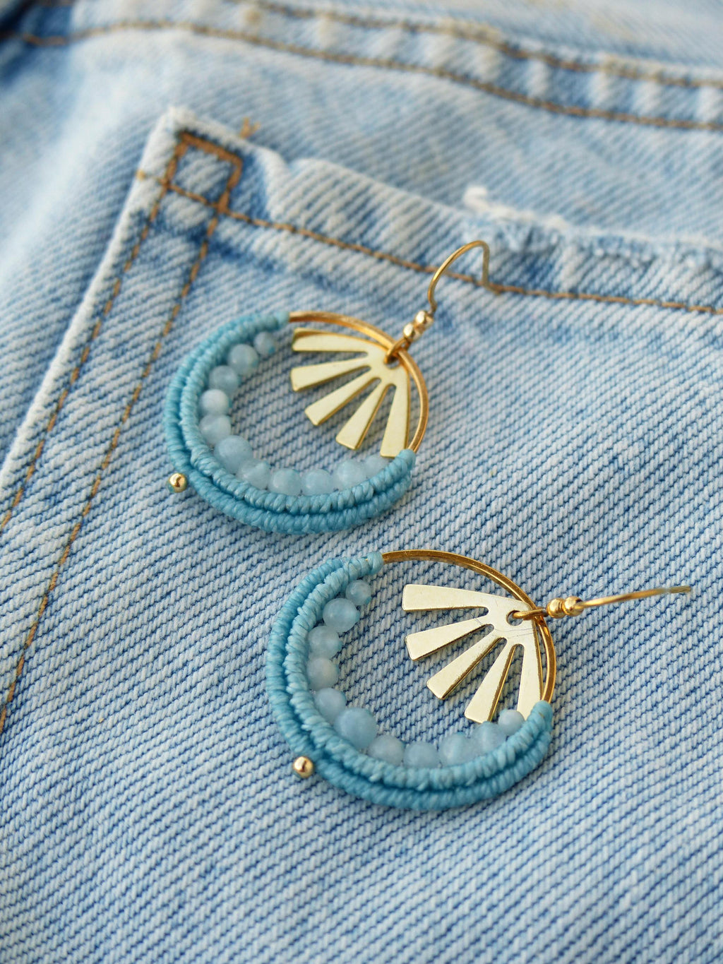 Peut inclure: Une paire de boucles d'oreilles faites à la main avec un motif de soleil. Les boucles d'oreilles présentent un charme doré en forme de soleil et un cercle de perles bleu clair. Elles sont posées sur un fond en jean bleu clair.
