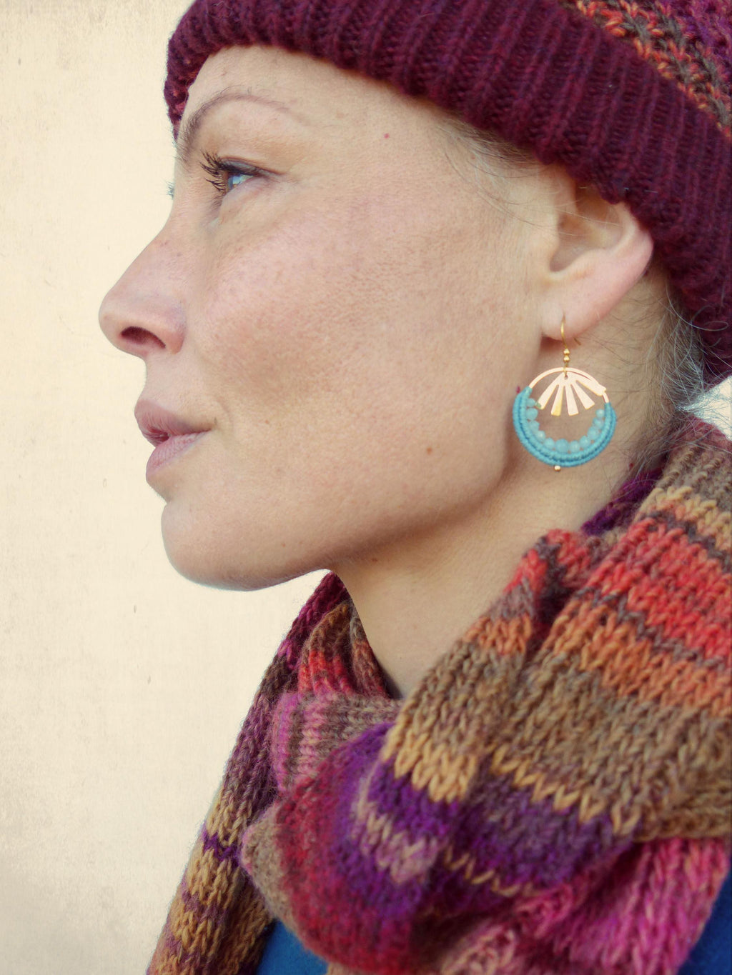 Peut inclure: Une femme porte des boucles d'oreilles dorées avec un cercle de perles turquoise et un motif d'éventail doré. Elle porte un bonnet en tricot bordeaux et une écharpe rayée colorée aux tons violets, oranges et bruns.