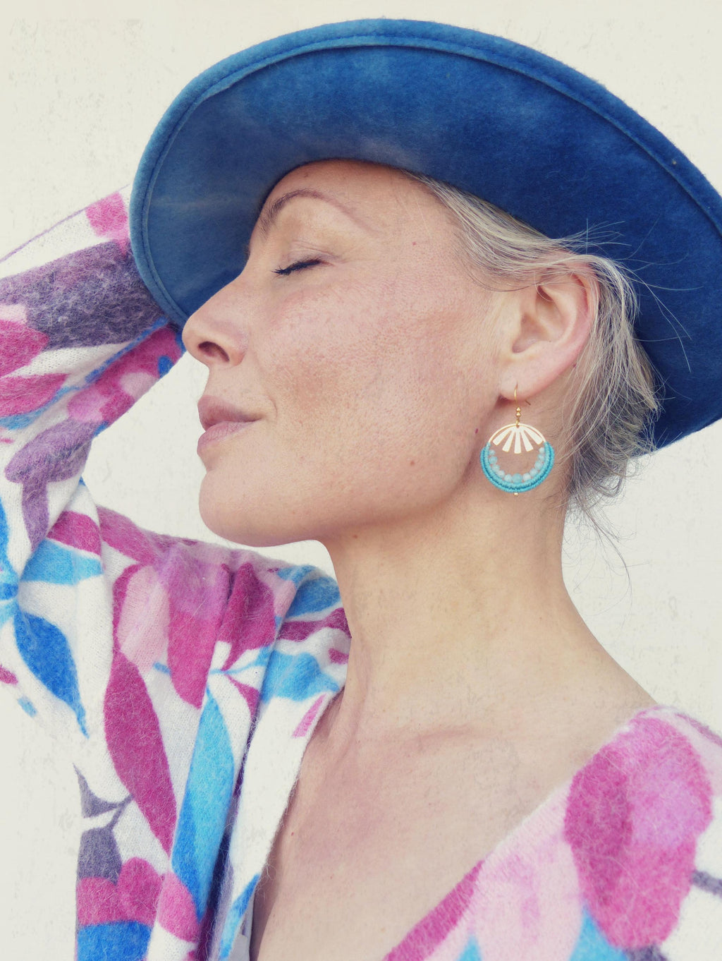Peut inclure: Une femme porte un chapeau bleu et des boucles d'oreilles. Les boucles d'oreilles sont dorées avec un motif turquoise et blanc. Elle porte un pull à motifs roses, bleus et violets.