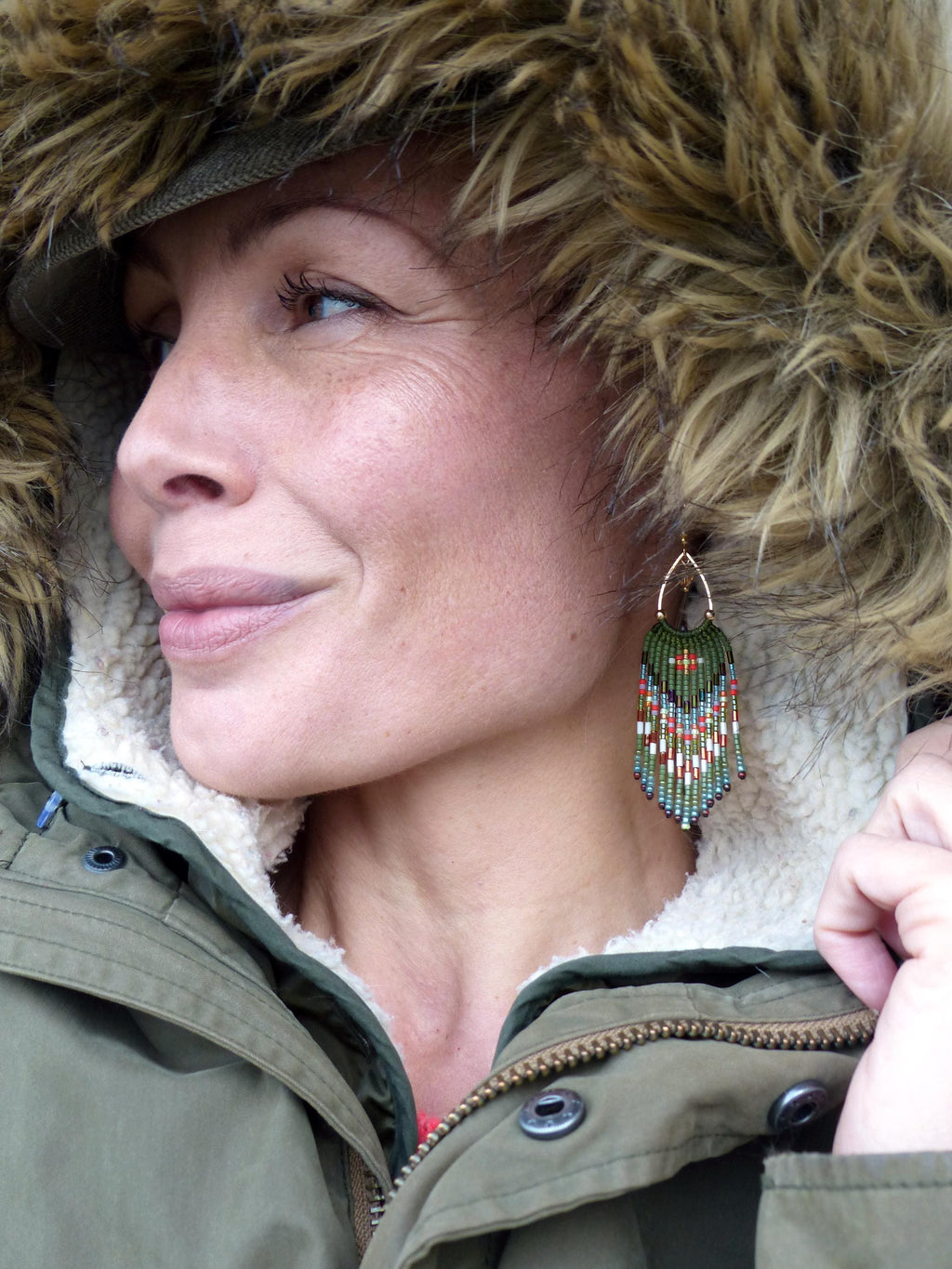 Peut inclure: Une femme porte une parka verte avec une capuche doublée de fourrure. Elle porte des boucles d'oreilles en perles avec un cerceau doré et un motif de perles vertes, rouges, blanches et bleues. Les boucles d'oreilles pendent de son oreille.