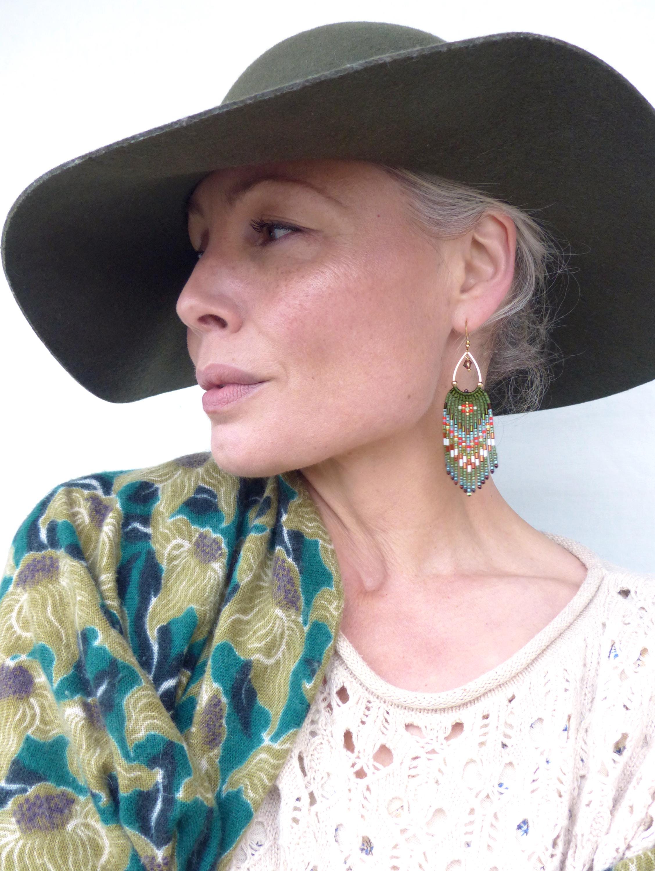 Peut inclure: Une femme porte un chapeau vert olive à larges bords et des boucles d'oreilles perlées. Les boucles d'oreilles ont un crochet doré et une frange de perles vertes, rouges et bleues. Une écharpe à motifs verts et crème est drapée sur les épaules.