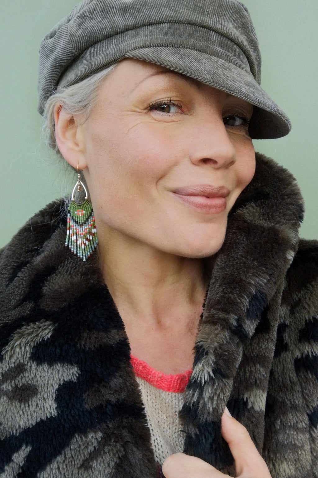 Peut inclure: Une femme porte une casquette grise et des boucles d'oreilles perlées. Les boucles d'oreilles ont un cadre en métal argenté avec des franges vertes, roses et blanches. Elle porte un manteau en fausse fourrure avec un motif camouflage.