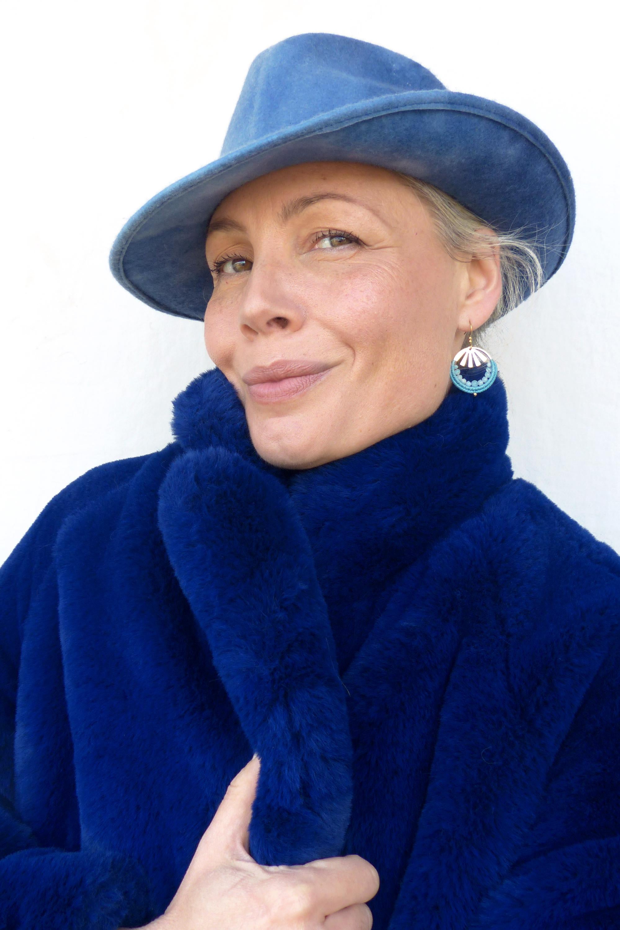 Peut inclure: Une femme porte un chapeau fedora bleu, un manteau en fausse fourrure bleu cobalt et des boucles d'oreilles turquoise et argentées. Le manteau a un col haut et le chapeau a un large bord. Le fond est un mur blanc uni.