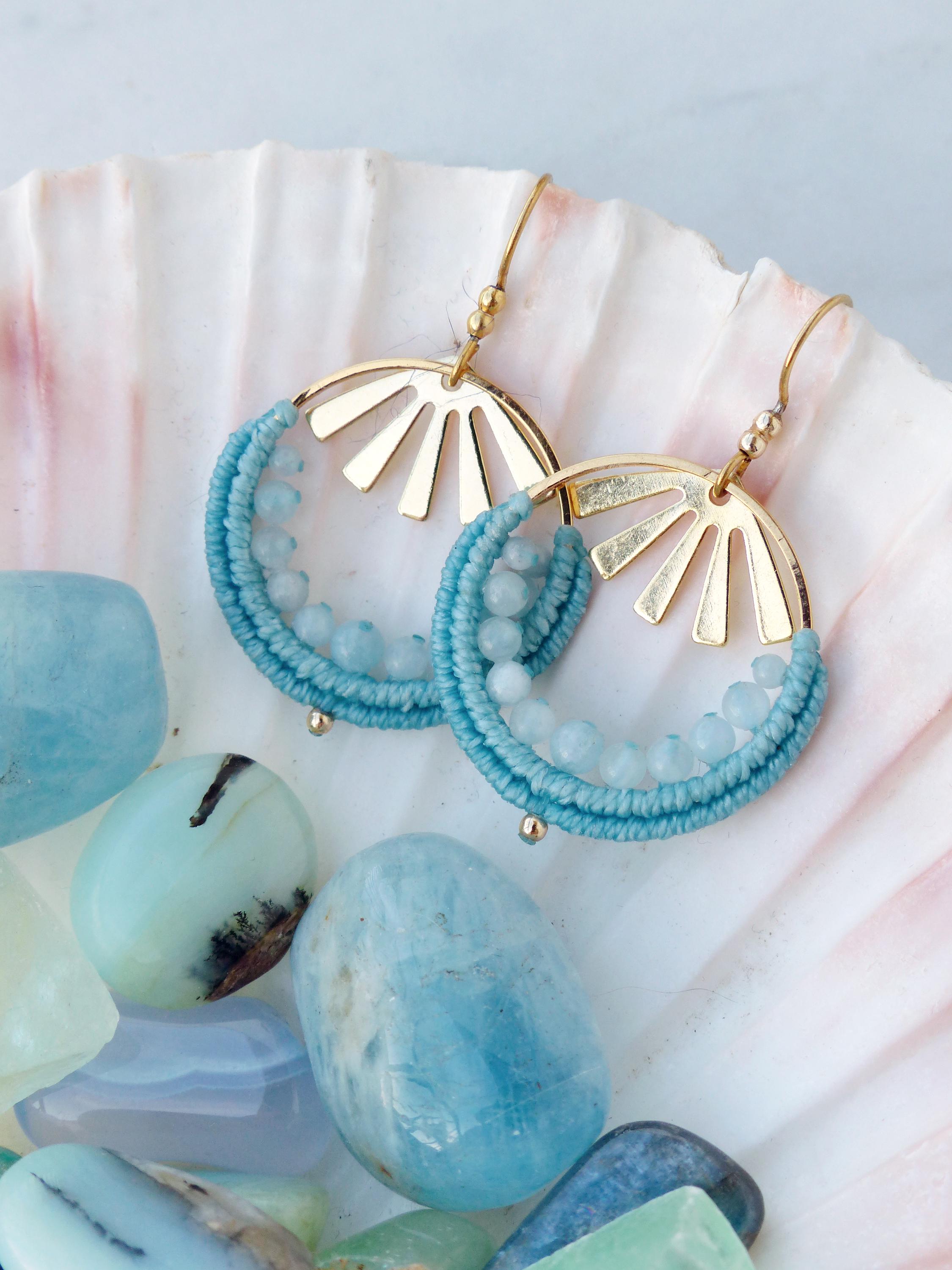 Peut inclure: Une paire de boucles d'oreilles dorées en forme de soleil, avec un design en macramé turquoise et de petites perles bleu clair. Les boucles d'oreilles sont présentées sur un coquillage blanc, entourées de pierres bleues et turquoise.