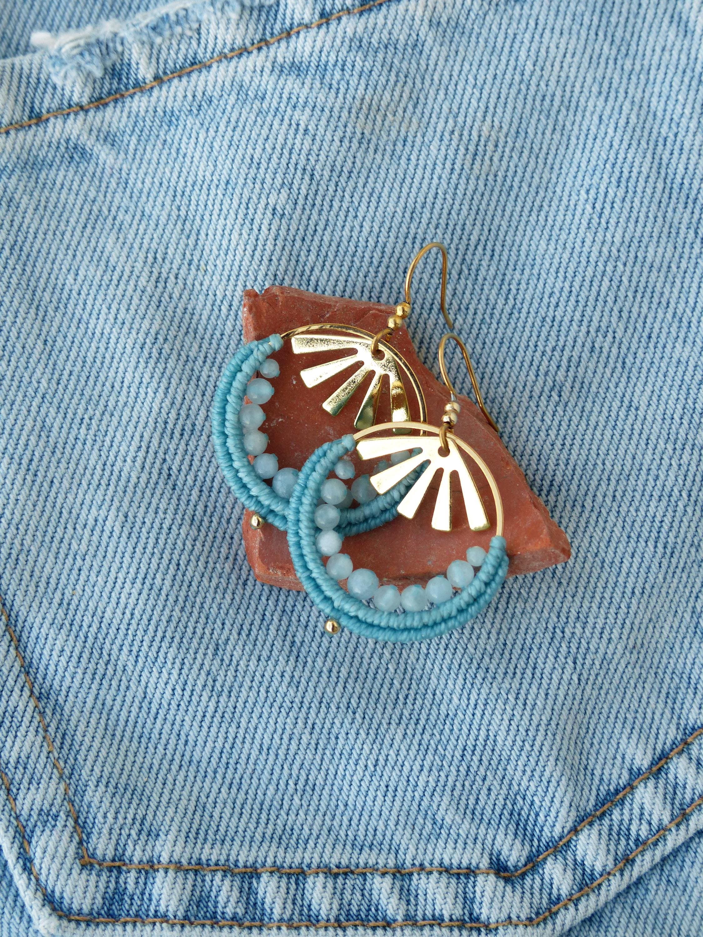 Peut inclure: Boucles d'oreilles dorées en forme de cerceau avec un motif de soleil. Les cerceaux sont enveloppés d'un cordon bleu clair et ornés de petites perles bleu clair. Les boucles d'oreilles reposent sur une petite pierre brun-rougeâtre, sur un fond en jean.