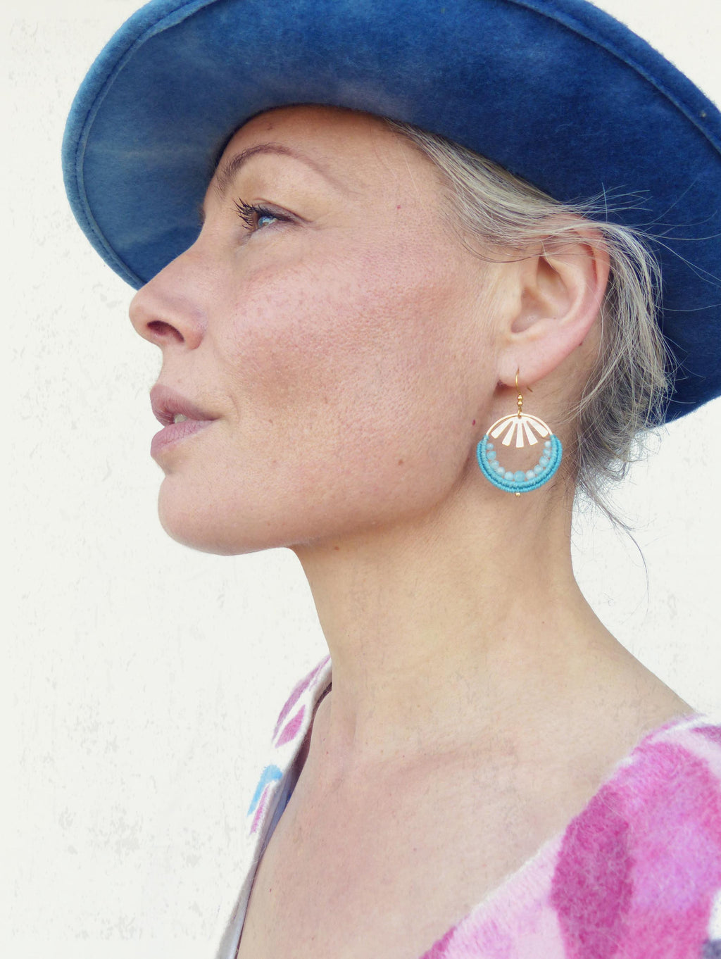 Peut inclure: Profil d'une personne portant un chapeau bleu et des boucles d'oreilles dorées. Les boucles d'oreilles ont un design circulaire avec un cadre doré, des perles turquoise et des accents blancs. La personne porte un haut à motifs roses et violets.