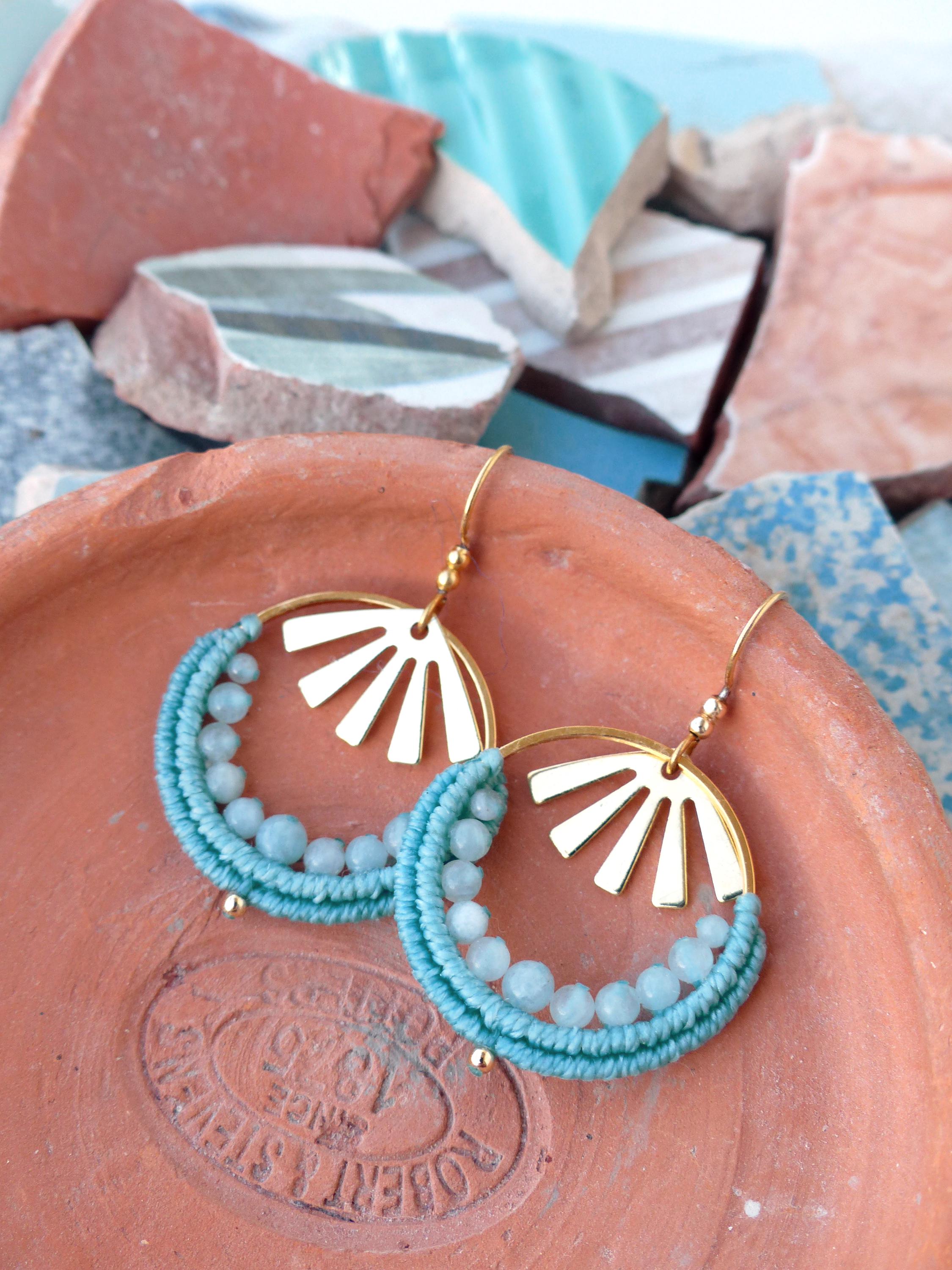 Peut inclure: Boucles d'oreilles dorées en forme de cerceau avec un motif de soleil. Les cerceaux sont ornés de macramé bleu clair et de petites perles rondes bleu clair. Les boucles d'oreilles sont présentées sur une surface couleur terre cuite.