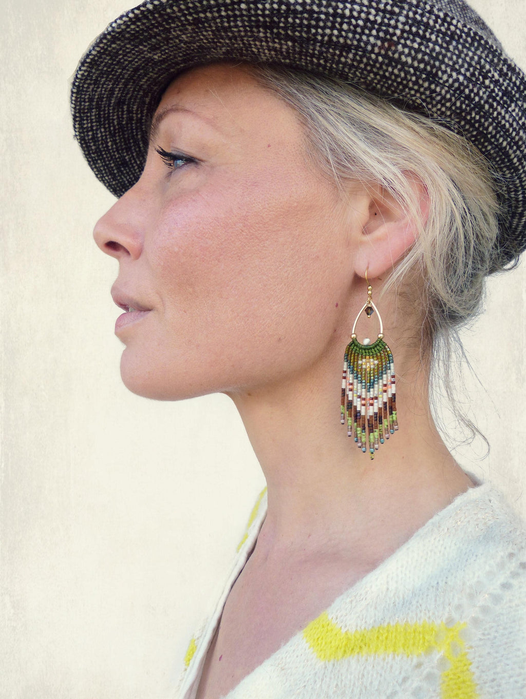 Peut inclure: Vue de profil d'une personne portant une casquette de style gavroche en tweed et un pull blanc à rayures jaunes. La personne porte une boucle d'oreille dorée en forme de goutte d'eau avec des franges de perles colorées.