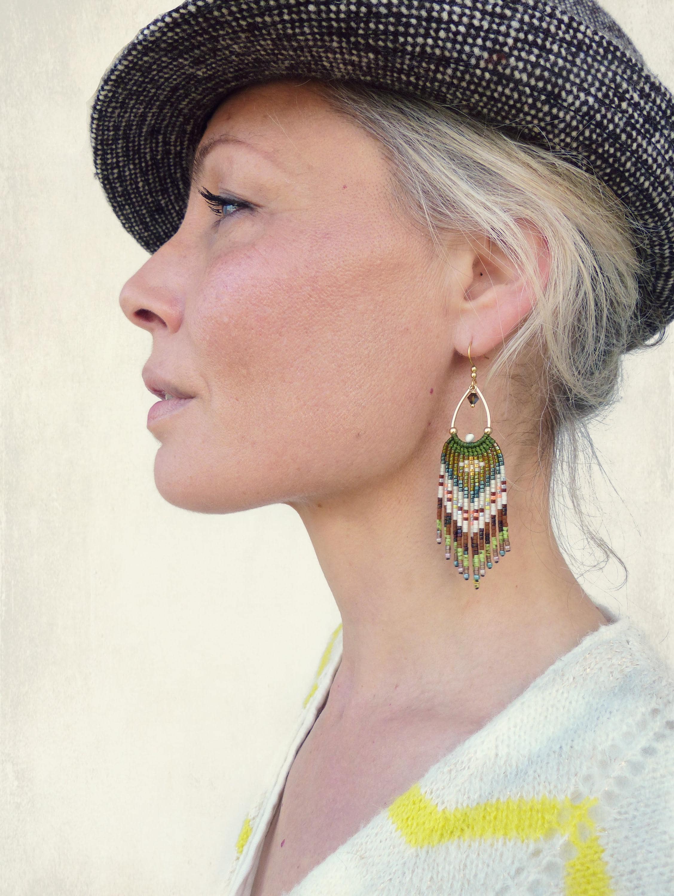 Peut inclure: Vue de profil d'une personne portant une casquette de style gavroche en tweed et un pull blanc à rayures jaunes. La personne porte une boucle d'oreille dorée en forme de goutte d'eau avec des franges de perles colorées.
