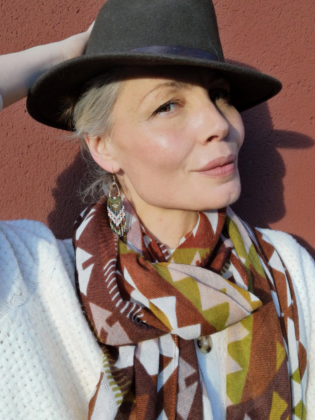 Peut inclure: Une femme portant un chapeau fedora gris foncé avec un ruban violet, une écharpe à motifs dans les tons marron, blanc et vert, et un pull blanc. La femme porte également des boucles d'oreilles.