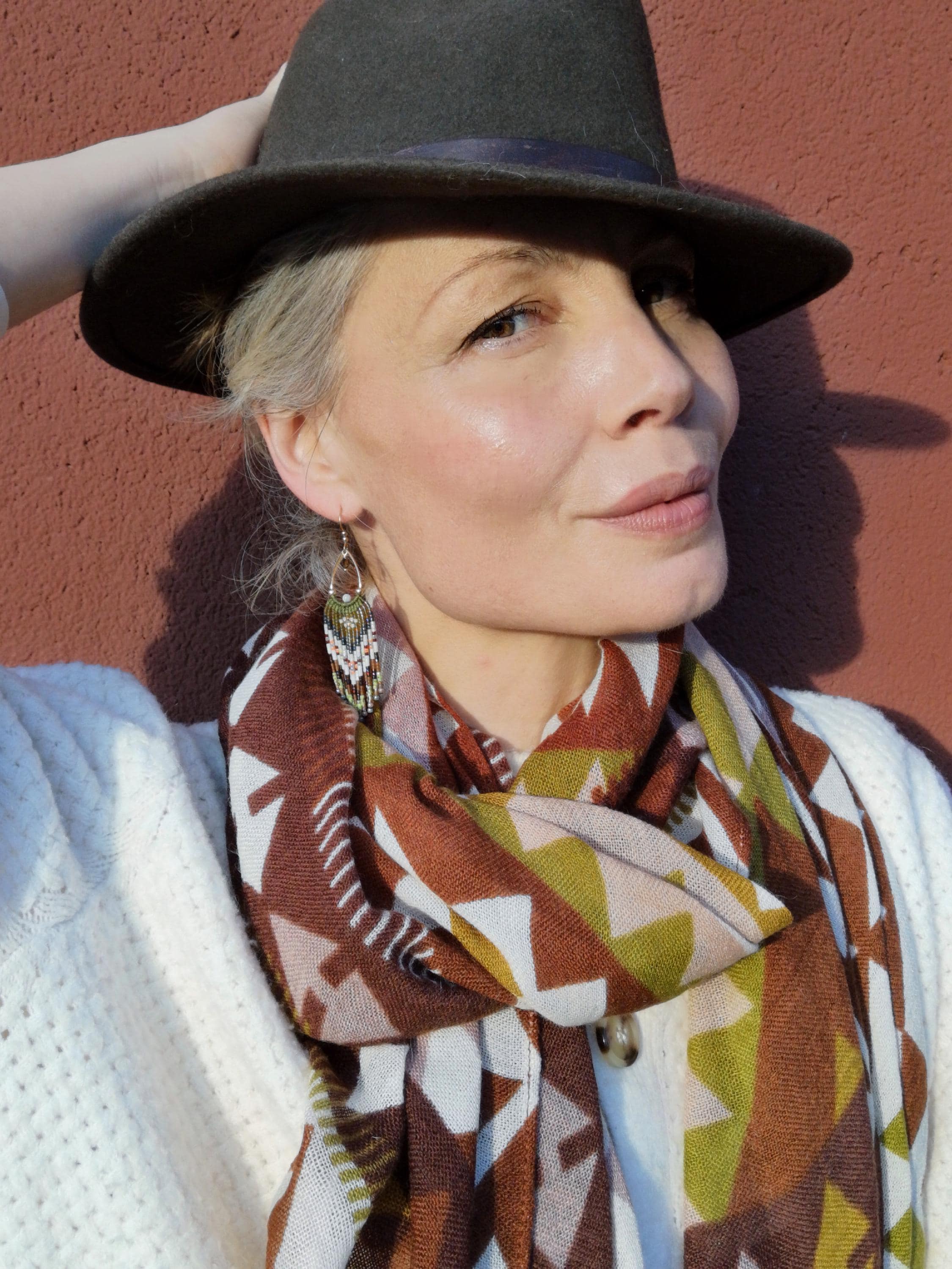 Peut inclure: Une femme portant un chapeau fedora gris foncé avec un ruban violet, une écharpe à motifs dans les tons marron, blanc et vert, et un pull blanc. La femme porte également des boucles d'oreilles.