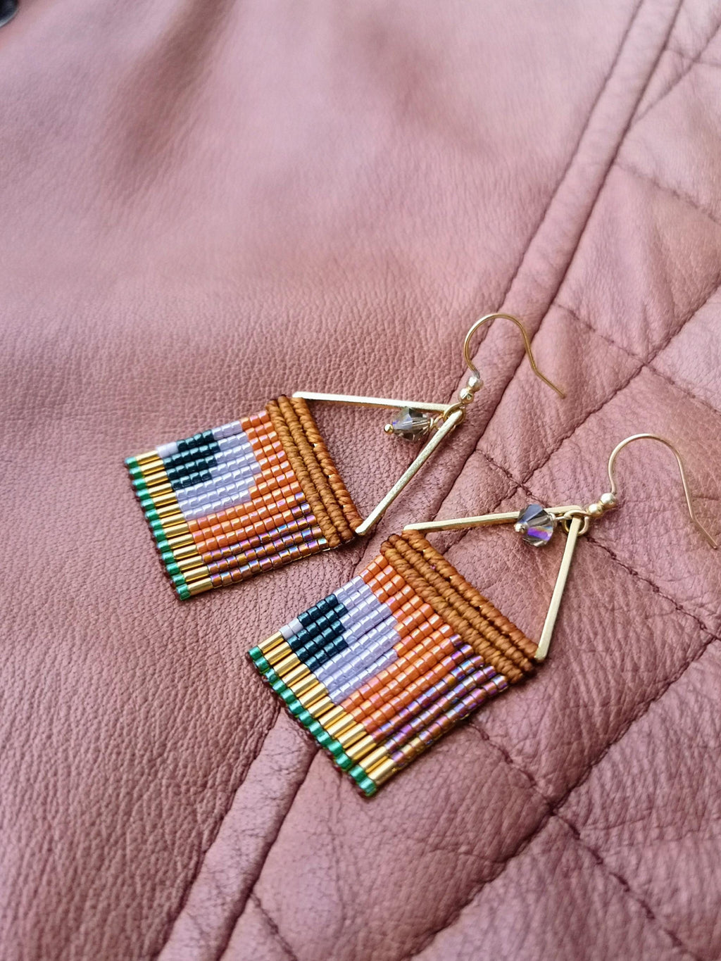 Peut inclure: Une paire de boucles d'oreilles faites à la main avec un design géométrique. Les boucles d'oreilles présentent un carré de perles colorées dans des tons orange, violet, vert et noir, fixées à un cadre triangulaire doré. De petits cristaux irisés sont attachés au cadre.