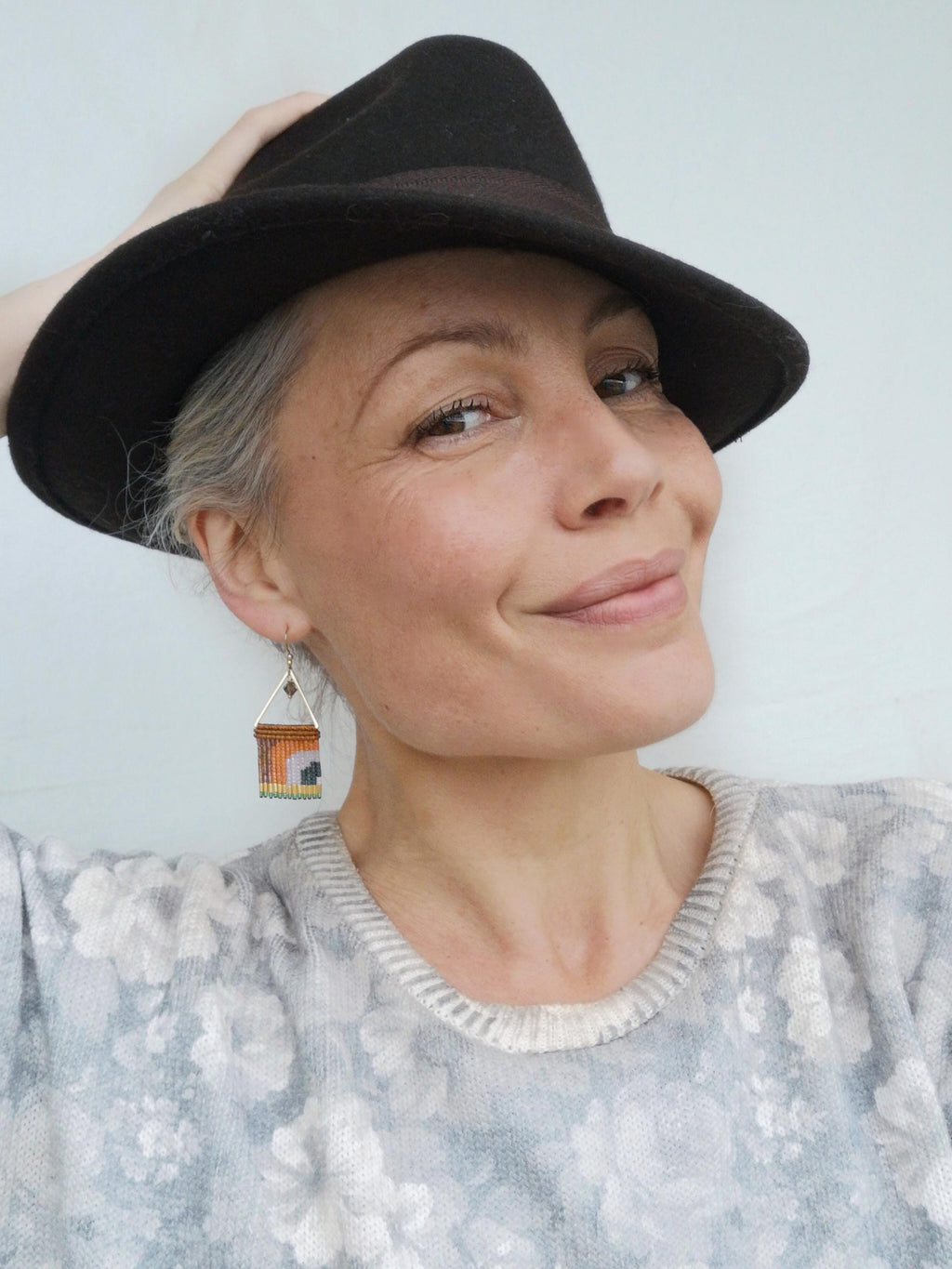 Peut inclure: Un chapeau fedora marron foncé avec une bande marron. La personne porte un pull gris à motifs floraux et des boucles d'oreilles en forme de maison perlées avec des détails orange, jaune et noir.
