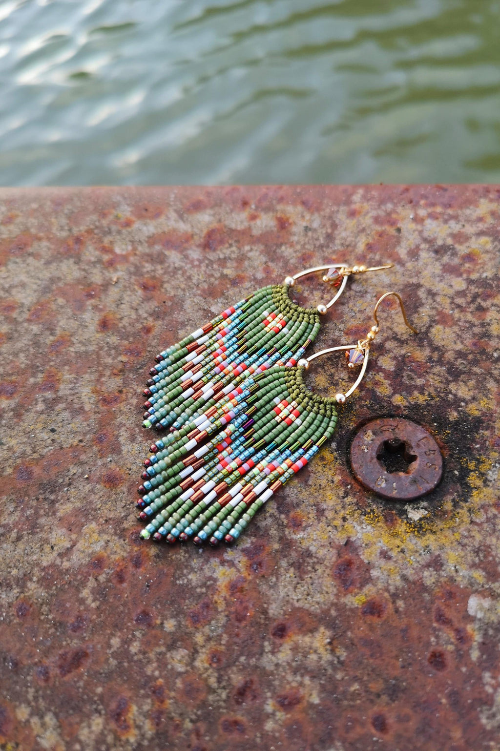 Peut inclure: Une paire de boucles d'oreilles en perles avec des crochets dorés. Les boucles d'oreilles présentent un motif de franges avec des perles vertes, rouges, blanches et bleues. Les boucles d'oreilles sont exposées sur une surface métallique rouillée.