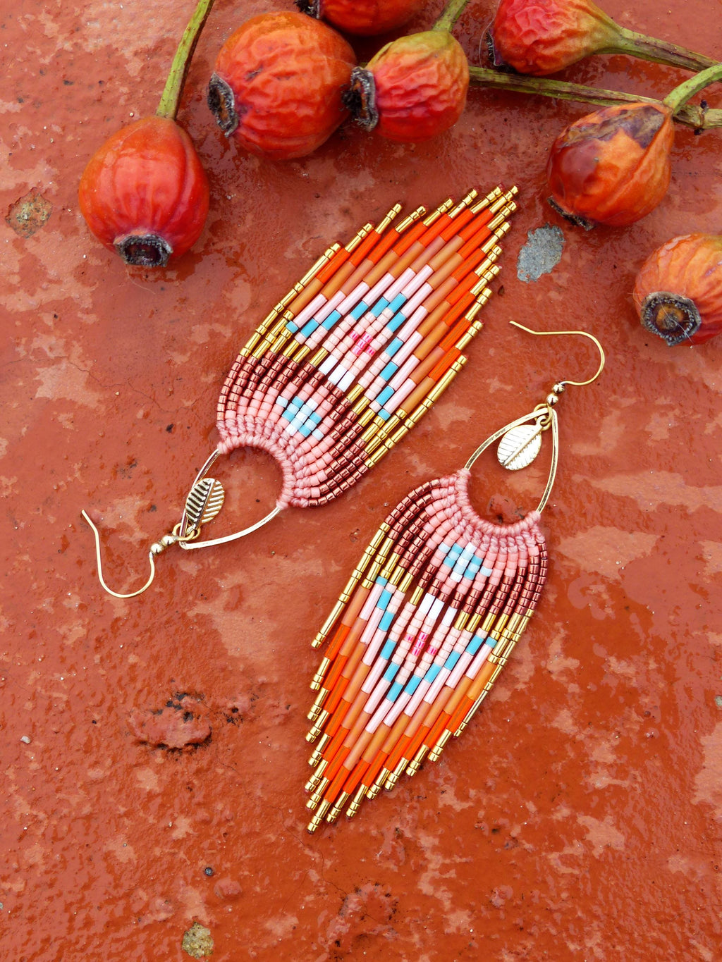 Peut inclure: Paire de boucles d'oreilles faites à la main en perles orange, rose et bleu. Les boucles d'oreilles ont un design à franges avec des accents dorés et un crochet doré. Elles sont présentées sur une surface rouge-orange, avec des cynorhodons oranges en arrière-plan.