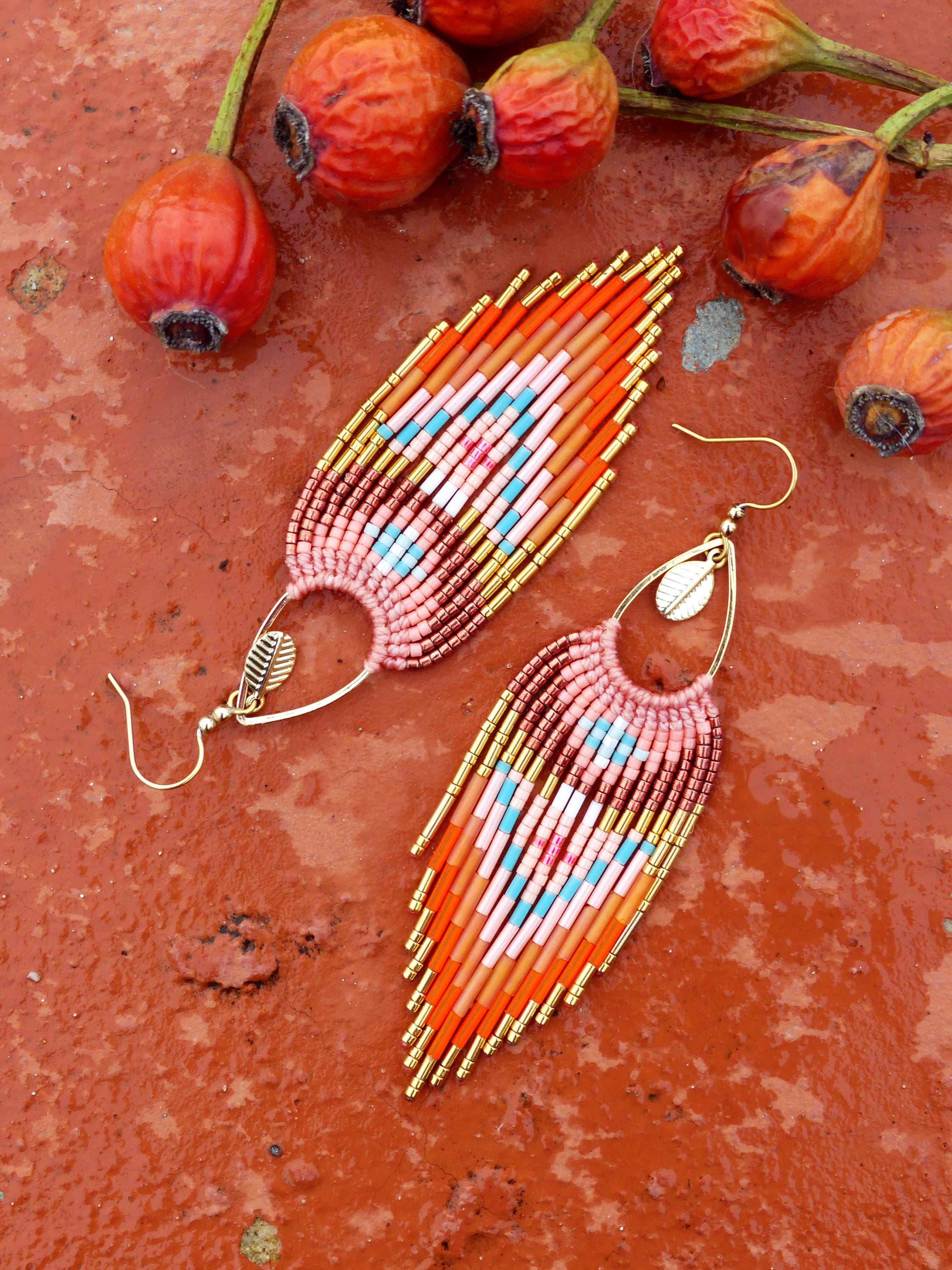 Peut inclure: Paire de boucles d'oreilles faites à la main en perles orange, rose et bleu. Les boucles d'oreilles ont un design à franges avec des accents dorés et un crochet doré. Elles sont présentées sur une surface rouge-orange, avec des cynorhodons oranges en arrière-plan.