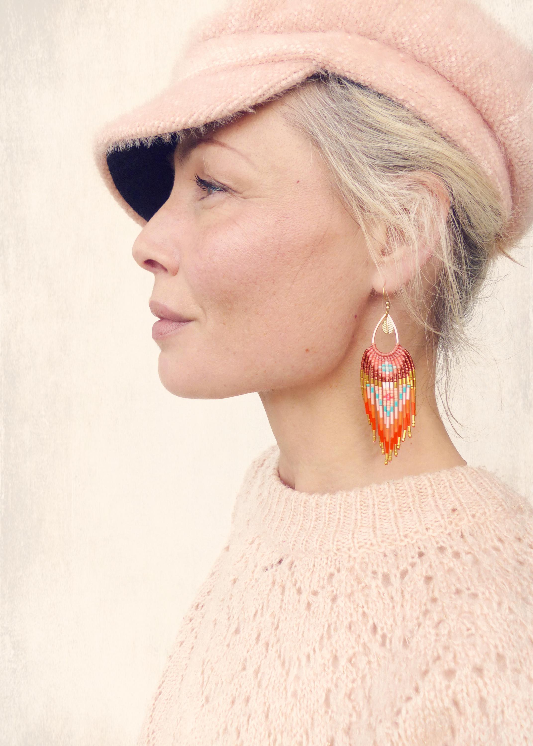 Peut inclure: Une femme porte un pull rose en tricot et un chapeau rose assorti. Elle porte des boucles d'oreilles en perles colorées avec des accents orange, bleus et jaunes. Les boucles d'oreilles ont une forme de goutte.