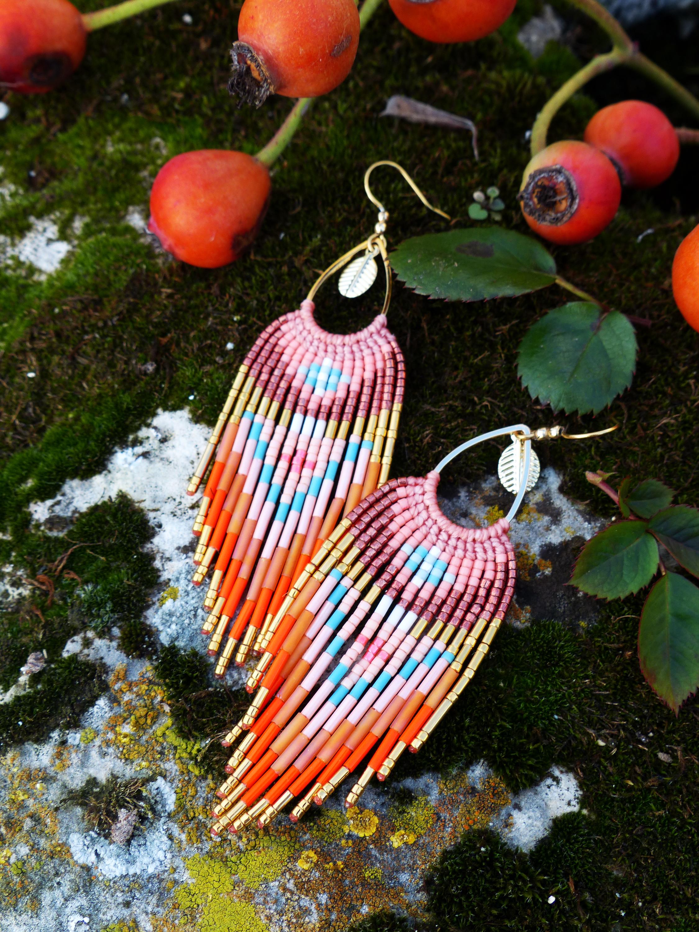 Peut inclure: Boucles d'oreilles faites à la main avec un design à franges. Elles présentent un dégradé de rose, orange, bleu et or. Les boucles sont suspendues à des crochets dorés avec une feuille décorative. Le fond montre des baies rouges et du feuillage vert.