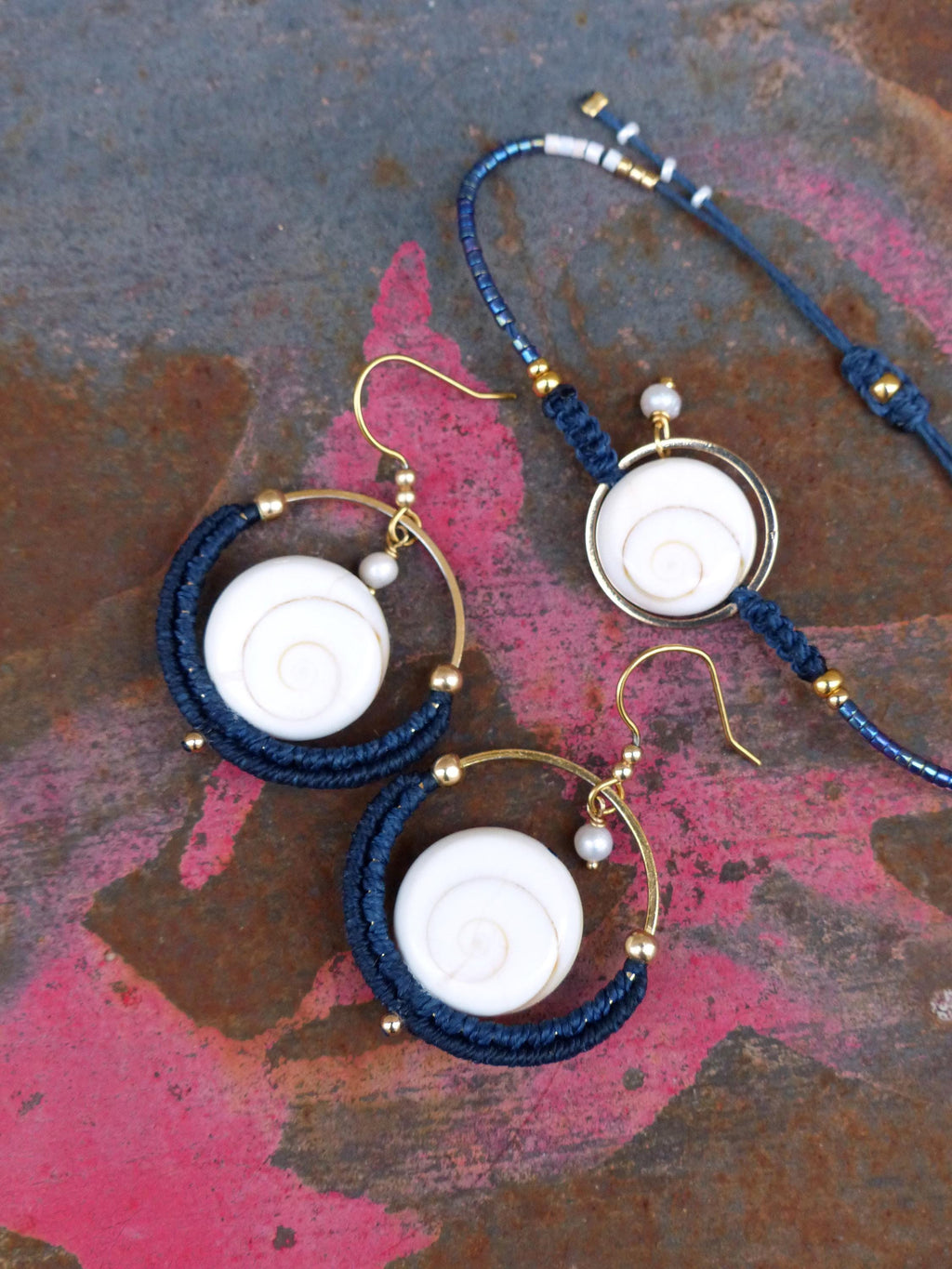 Peut inclure: Un ensemble de bijoux comprenant une paire de boucles d'oreilles créoles et un bracelet. Les boucles d'oreilles et le bracelet ont chacun un centre blanc en forme de spirale, entouré d'un motif tissé bleu marine. Des accents et crochets en métal doré complètent l'ensemble.