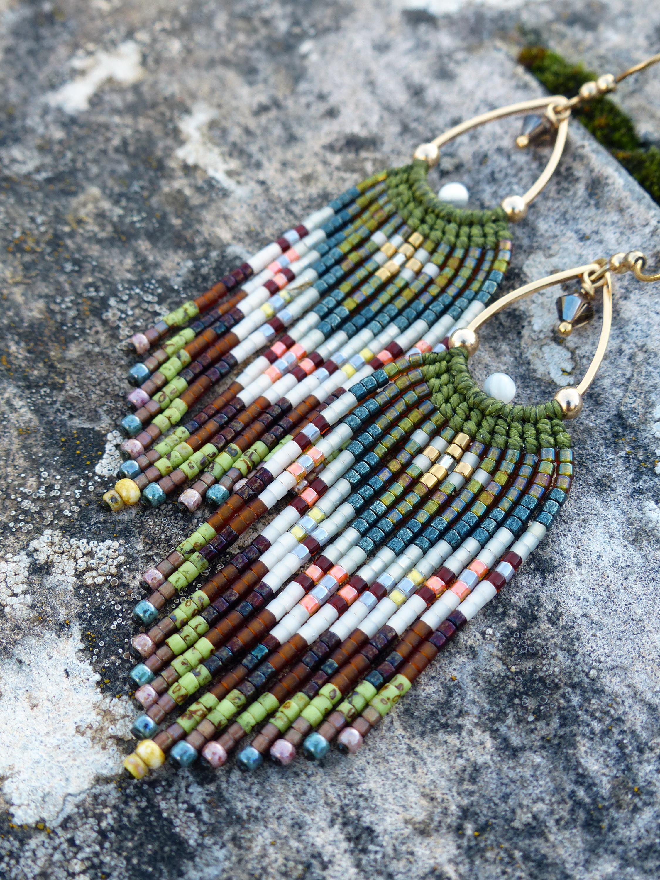 Peut inclure: Boucles d'oreilles faites à la main avec un design à franges. Les boucles d'oreilles présentent des crochets en métal doré et sont ornées de petites perles colorées dans les tons de vert, marron, blanc et bleu. Les boucles d'oreilles sont présentées sur une surface grise texturée.