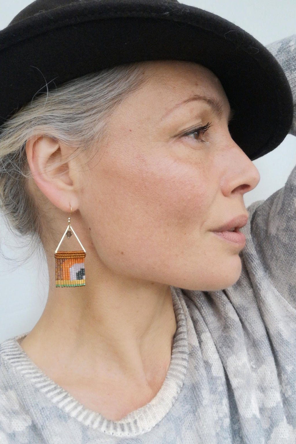 Peut inclure: Gros plan d'une personne portant un chapeau noir et un pull gris. L'attention se porte sur une paire de boucles d'oreilles faites à la main. Les boucles d'oreilles présentent un motif géométrique avec des perles orange, marron et jaunes, suspendues à des cadres triangulaires dorés.