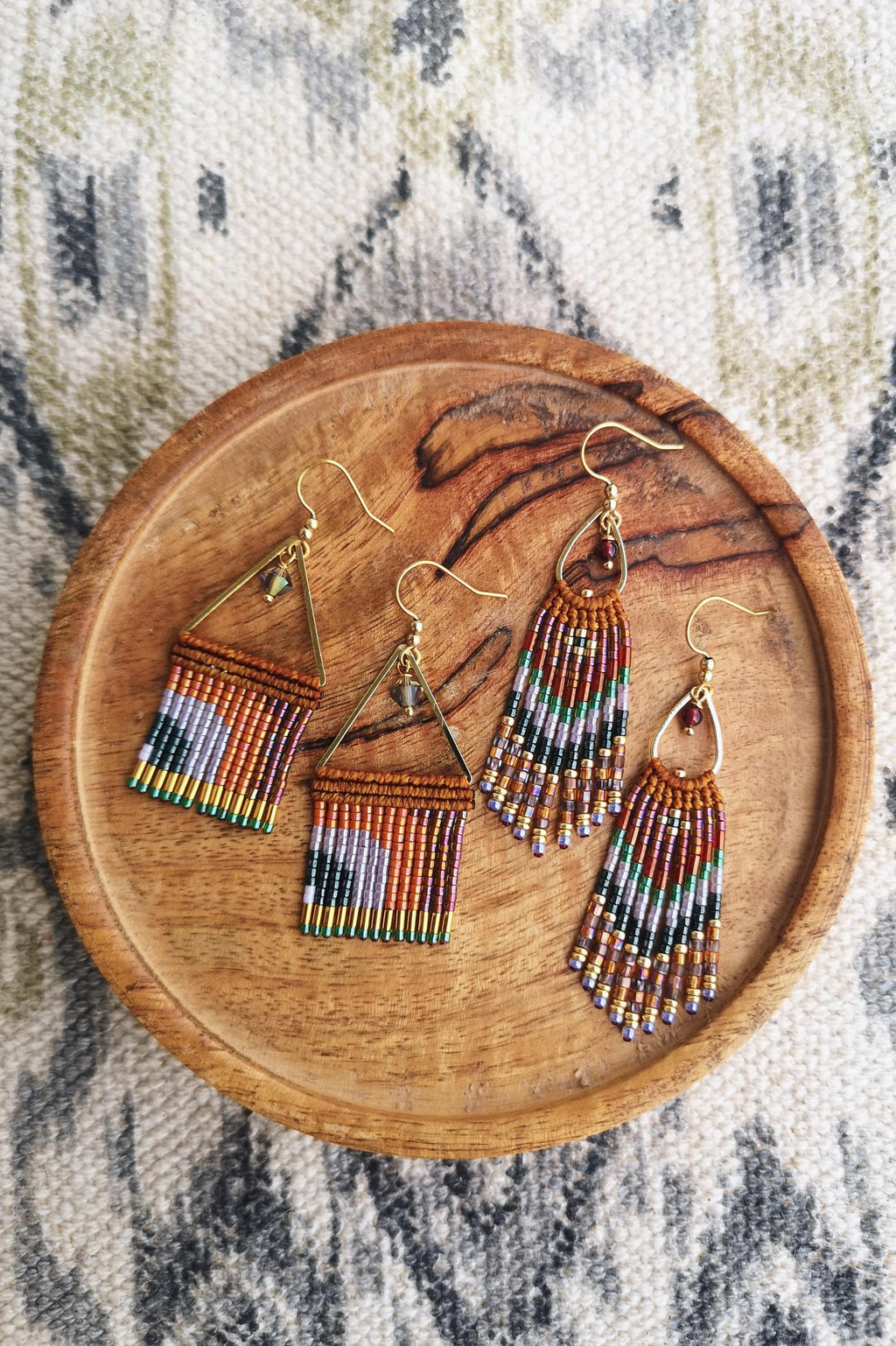 Peut inclure: Quatre paires de boucles d'oreilles perlées faites à la main de différentes formes et designs, présentées sur une assiette en bois. Les boucles d'oreilles comportent des crochets dorés et des perles colorées dans les tons orange, violet, vert et noir. L'assiette est posée sur un tissu à motifs.