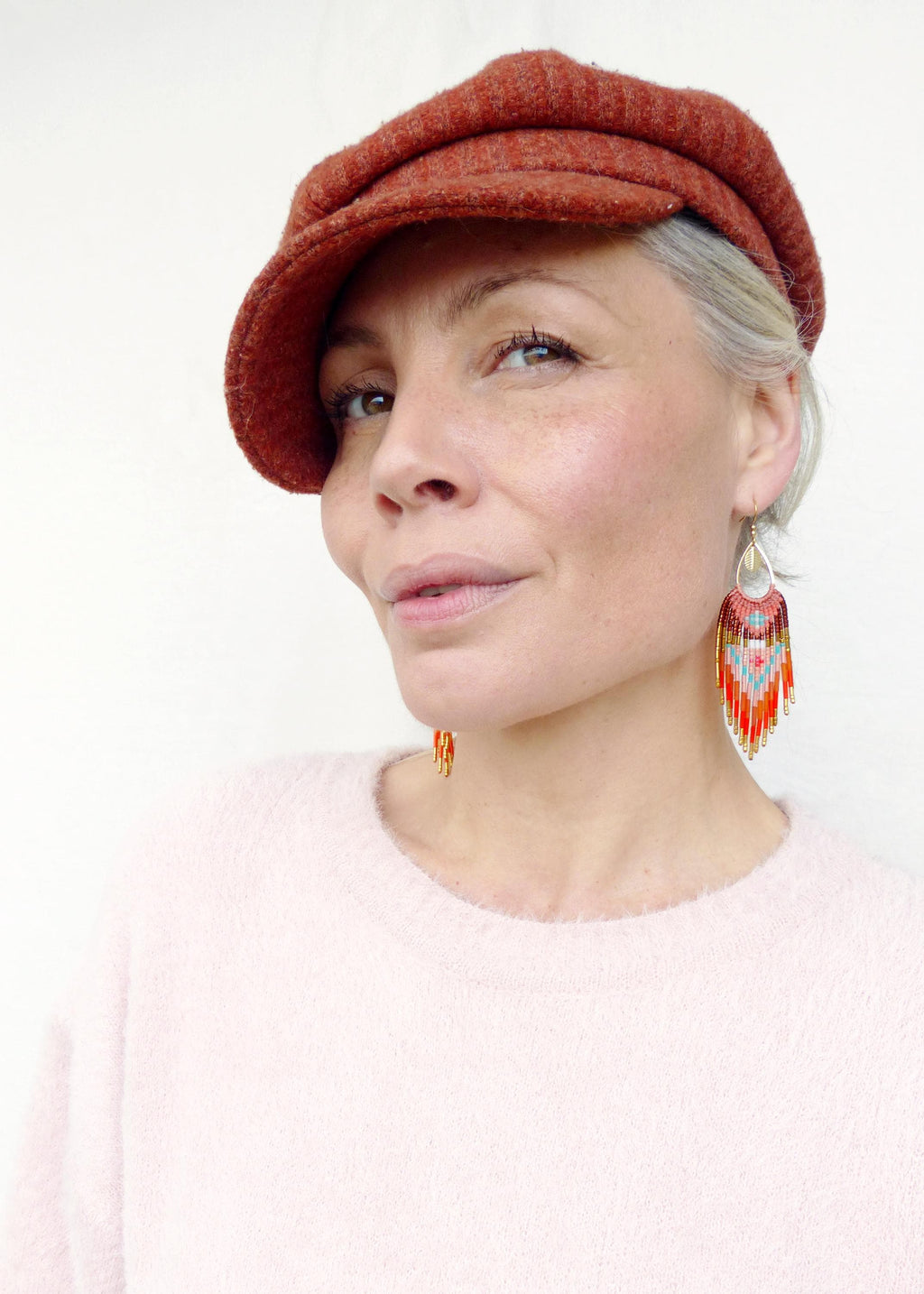 Peut inclure: Une femme porte une casquette de style gavroche rouille et un pull rose clair. Elle porte des boucles d'oreilles en perles avec une forme de goutte d'eau et un motif coloré de perles rouges, oranges, bleues et dorées. Les boucles d'oreilles sont une pièce maîtresse.