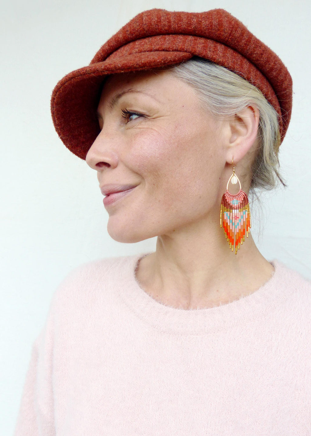 Peut inclure: Une femme porte une casquette de style gavroche rouille et un pull rose clair. Elle porte de longues boucles d'oreilles perlées avec un dessus doré et un motif de perles orange, roses et turquoise.