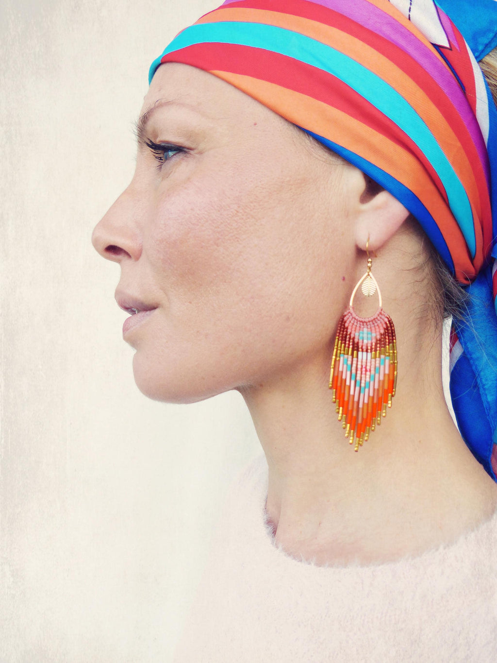 Peut inclure: Une femme porte un foulard coloré et des boucles d'oreilles en perles. Les boucles d'oreilles en forme de goutte d'eau ont un dessus doré et une frange de perles orange, rouges, blanches et bleues. Le foulard présente des rayures rouges, oranges, bleues et violettes.