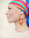 Peut inclure: Une femme porte un foulard coloré et des boucles d'oreilles en perles. Les boucles d'oreilles en forme de goutte d'eau ont un dessus doré et une frange de perles orange, rouges, blanches et bleues. Le foulard présente des rayures rouges, oranges, bleues et violettes.