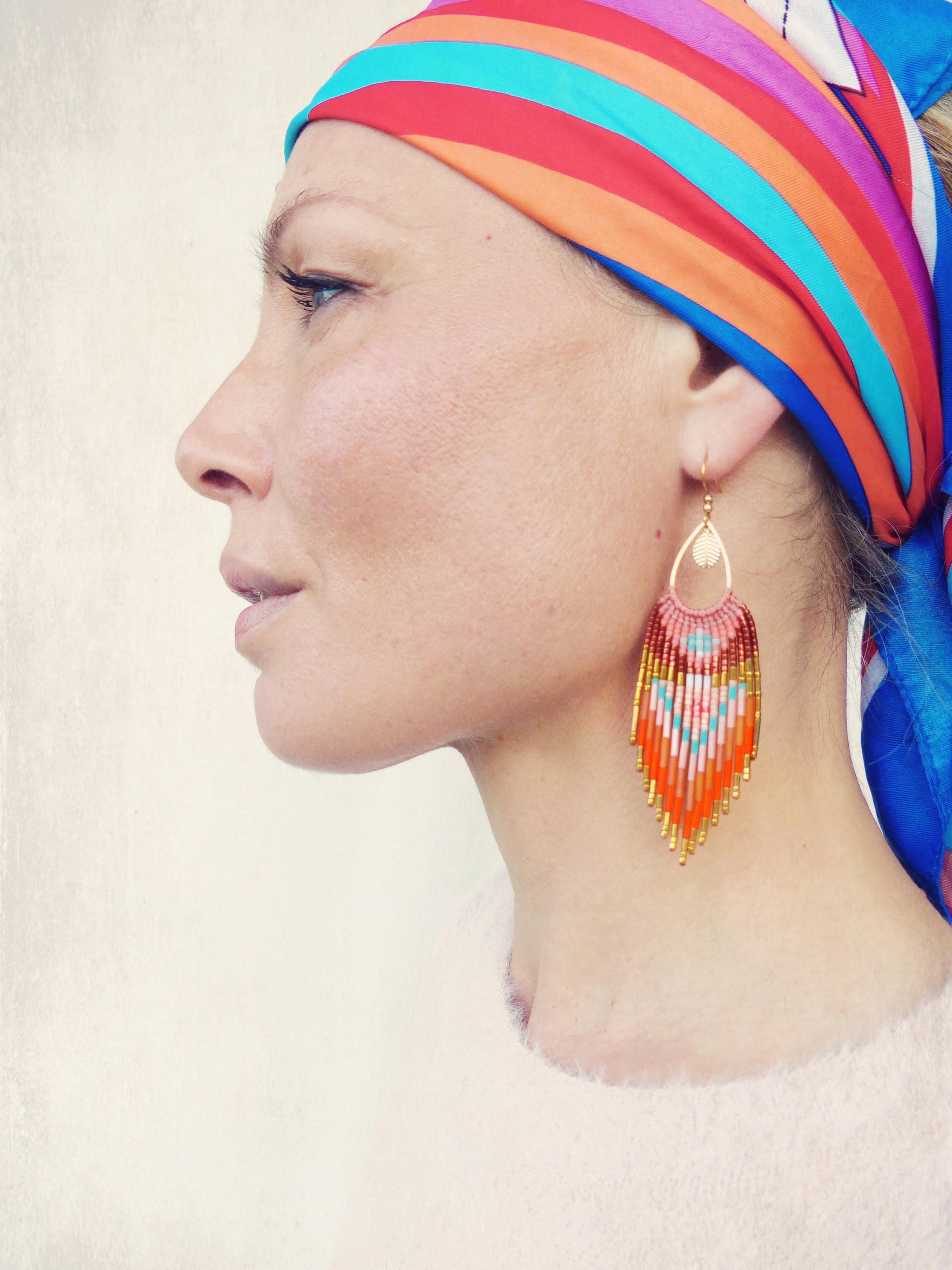 Peut inclure: Une femme porte un foulard coloré et des boucles d'oreilles en perles. Les boucles d'oreilles en forme de goutte d'eau ont un dessus doré et une frange de perles orange, rouges, blanches et bleues. Le foulard présente des rayures rouges, oranges, bleues et violettes.