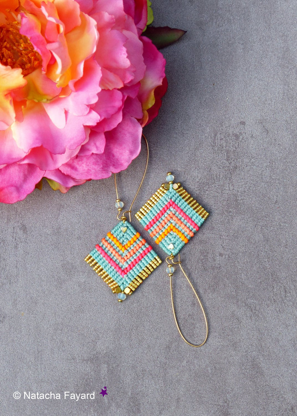 Peut inclure: Une paire de boucles d'oreilles pendantes dorées avec des perles géométriques turquoise, rose et orange. Les boucles d'oreilles sont sur un fond gris.