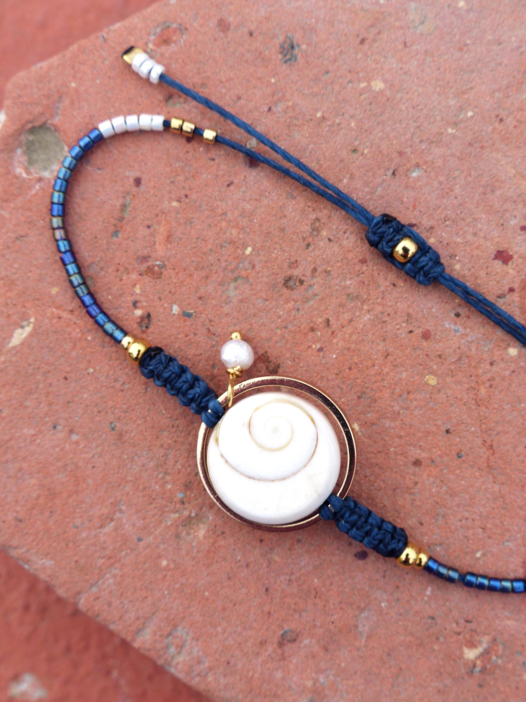 Peut inclure: Bracelet artisanal avec un pendentif en forme de coquillage blanc en spirale, serti dans un cadre doré. Le bracelet est composé d'un cordon bleu foncé avec des détails dorés et de petites perles bleues et argentées. Une petite perle blanche est attachée au pendentif.