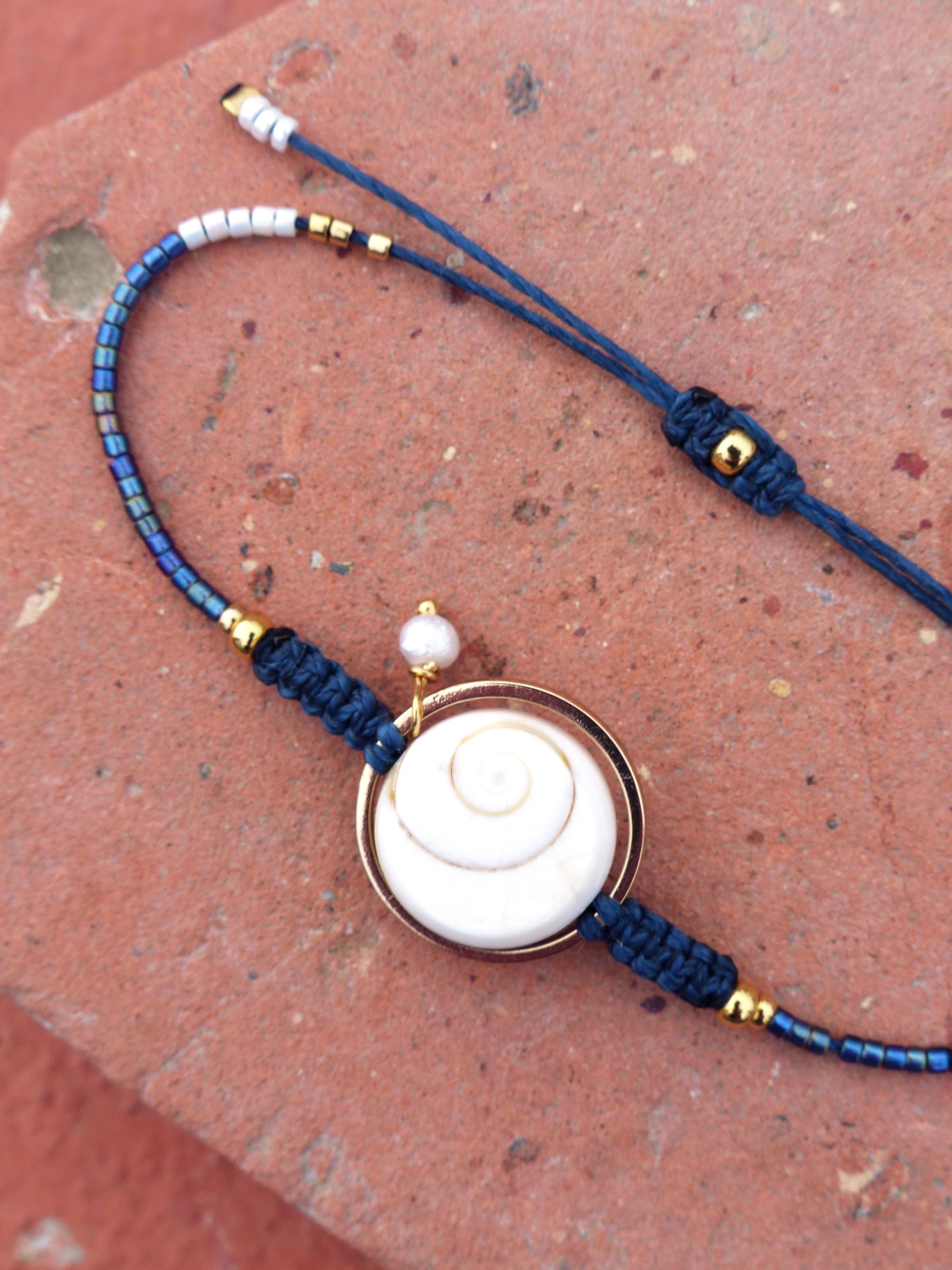 Peut inclure: Bracelet artisanal avec un pendentif en forme de coquillage blanc en spirale, serti dans un cadre doré. Le bracelet est composé d'un cordon bleu foncé avec des détails dorés et de petites perles bleues et argentées. Une petite perle blanche est attachée au pendentif.
