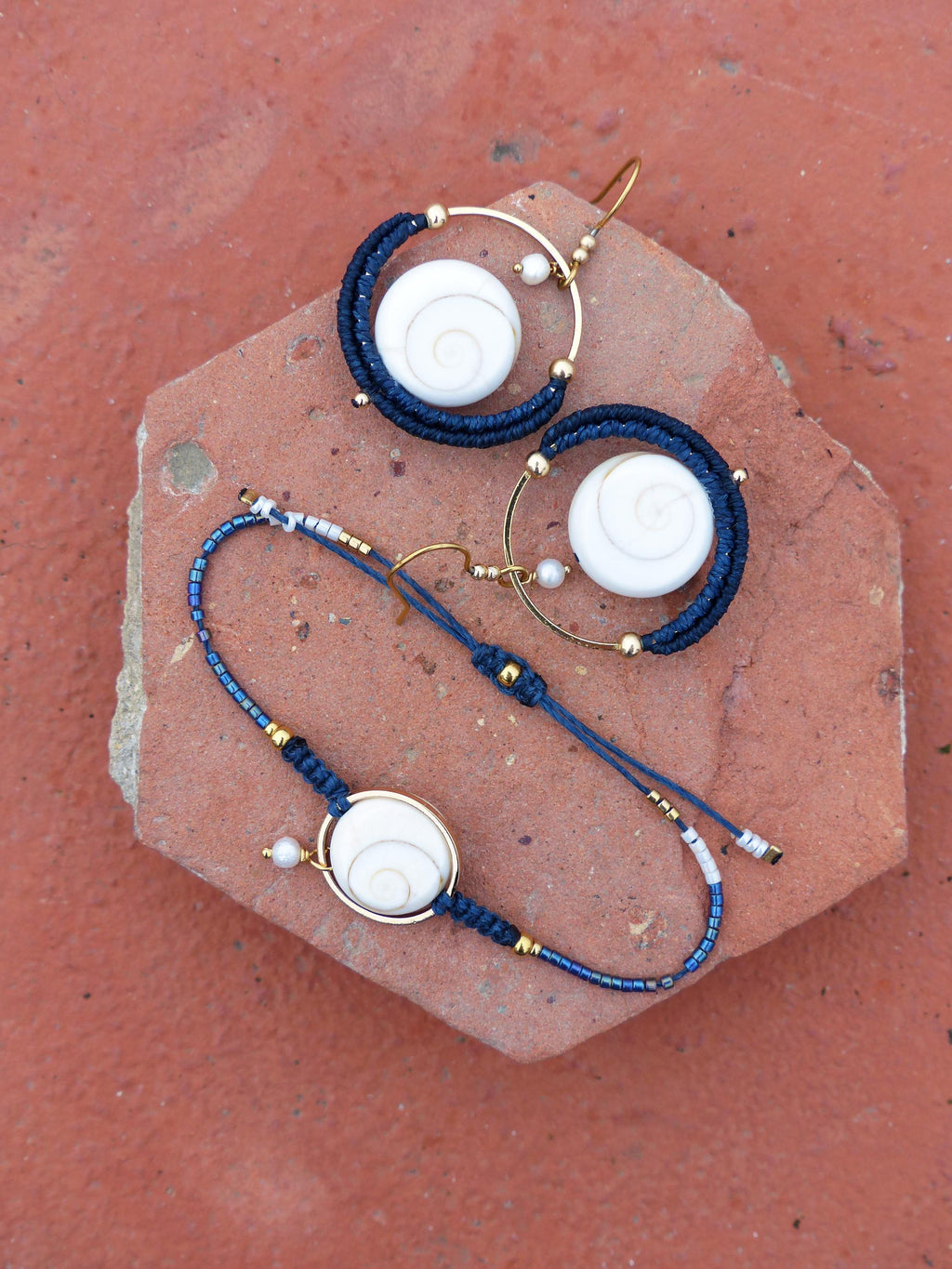 Peut inclure: Un ensemble de bijoux comprenant un bracelet et des boucles d'oreilles. Le bracelet et les boucles d'oreilles ont un cadre en métal doré avec un motif en spirale blanc. Les boucles d'oreilles sont de style créole avec un cordon bleu foncé. Le bracelet a un cordon bleu foncé et de petites perles.