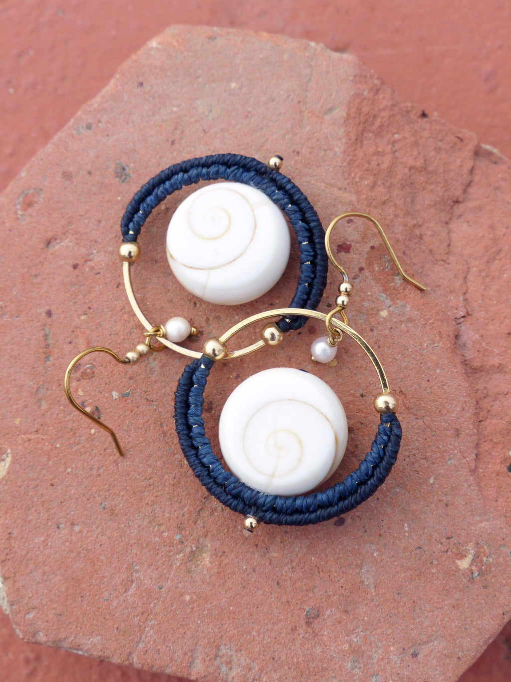 Peut inclure: Boucles d'oreilles faites à la main avec des perles blanches à motif en spirale, rehaussées de macramé bleu marine et de métal doré. De petites perles et des crochets dorés complètent le design. Présentées sur une surface brun-rouge.
