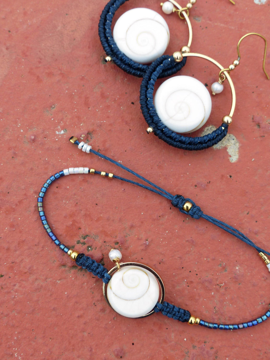 Peut inclure: Ensemble de bijoux faits à la main comprenant un bracelet et des boucles d'oreilles. Les boucles d'oreilles ont des cerceaux dorés avec un motif en spirale blanc et du macramé bleu marine. Le bracelet a un motif en spirale blanc similaire et du macramé bleu marine.