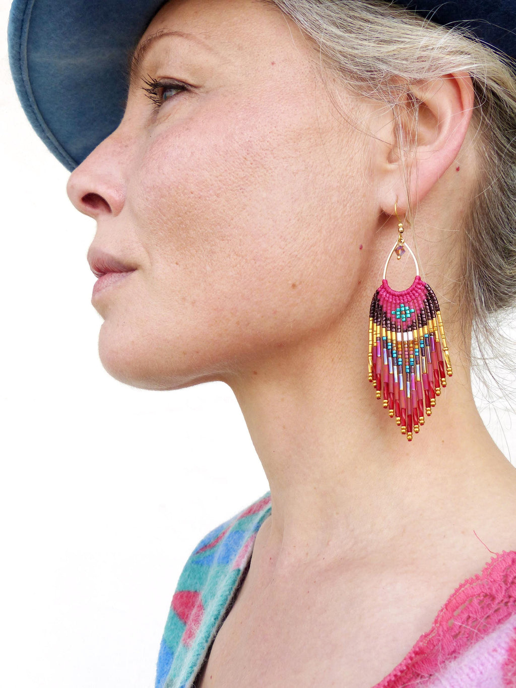 Peut inclure: Gros plan sur une boucle d'oreille perlée avec un motif rose, marron, doré et turquoise. La boucle d'oreille a un cerceau doré et une frange de perles colorées. La boucle d'oreille est portée par une personne aux cheveux gris et un chapeau bleu.