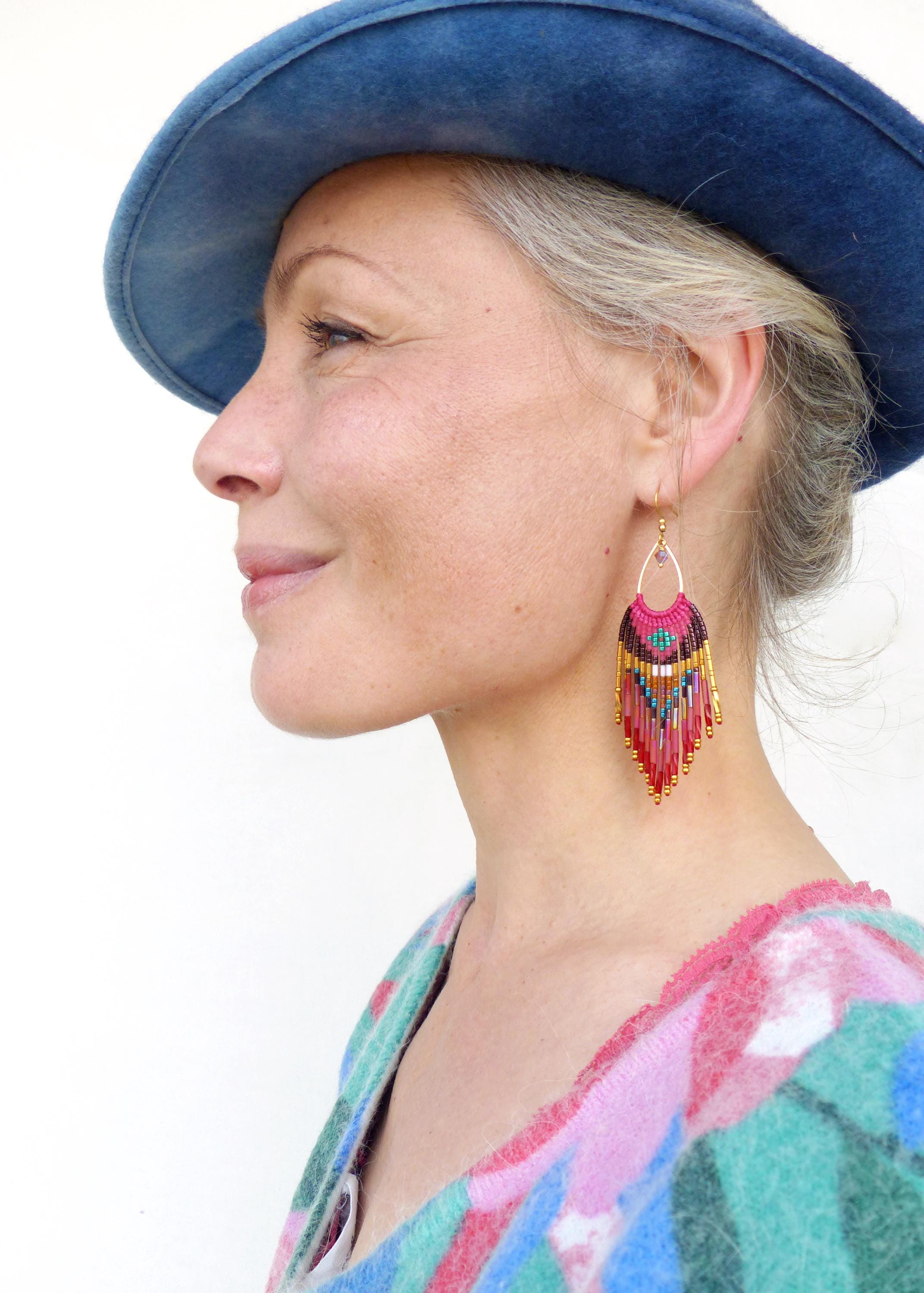 Peut inclure: Une femme porte des boucles d'oreilles en perles colorées avec un cerceau doré et un chapeau bleu. Les boucles d'oreilles présentent un code couleur rose, rouge et or avec un motif de franges. La femme porte un haut à motifs.