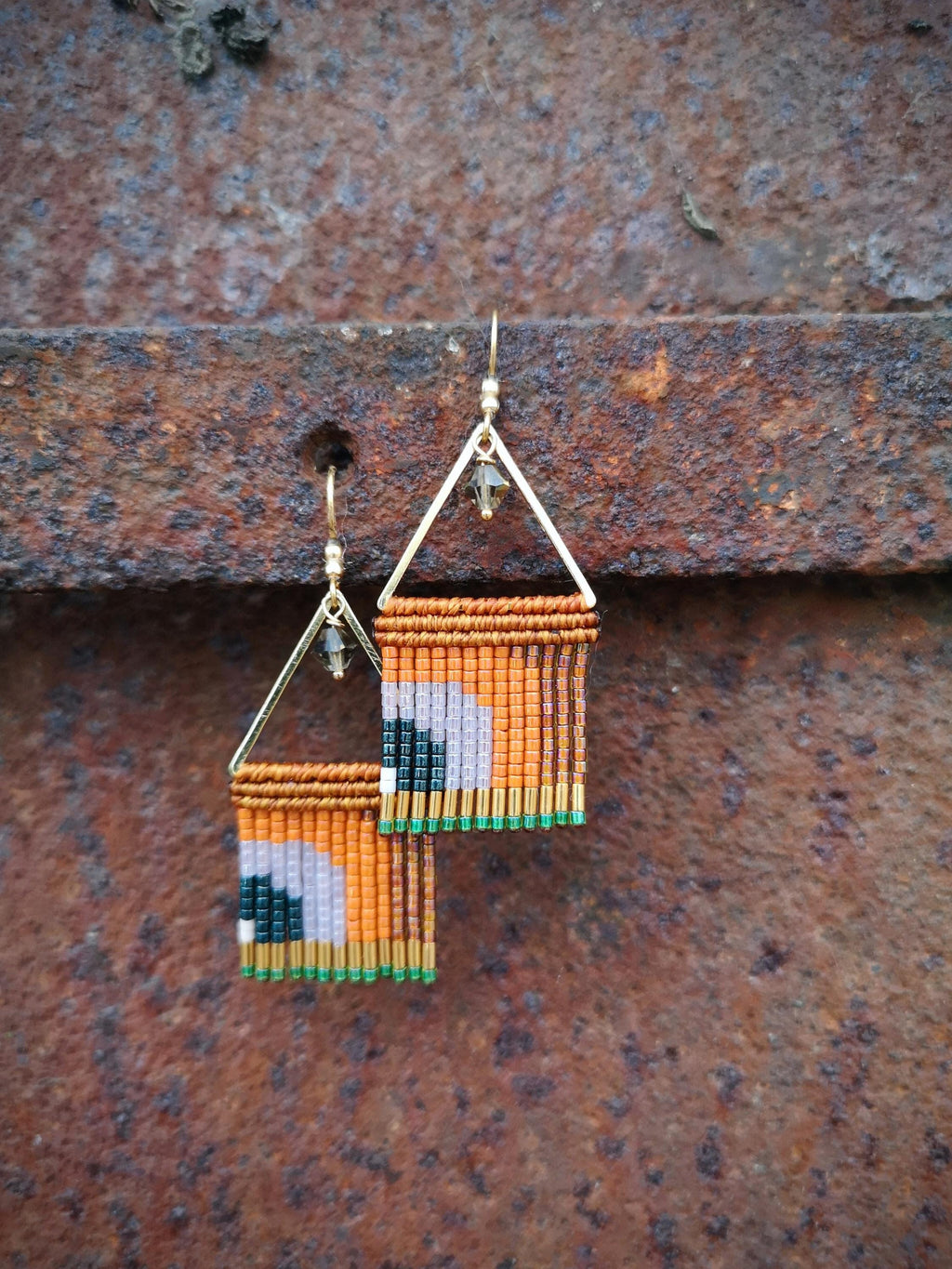 Peut inclure: Boucles d'oreilles faites à la main avec un design géométrique. Les boucles d'oreilles présentent un haut triangulaire doré et un bas rectangulaire avec des perles orange, noires, blanches et vertes. Elles sont suspendues sur un fond en métal rouillé.