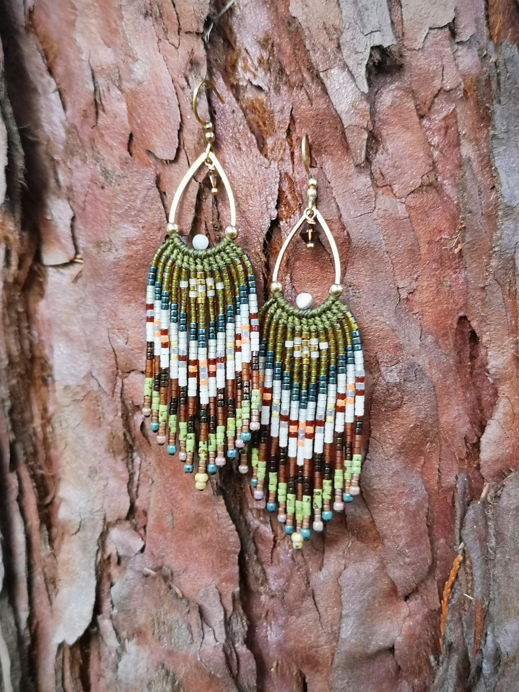 Peut inclure: Une paire de boucles d'oreilles faites à la main avec des perles et une forme de goutte d'eau. Les boucles d'oreilles ont des crochets en métal doré et une frange de perles colorées dans les tons de marron, vert, blanc et orange. Fond marron texturé.