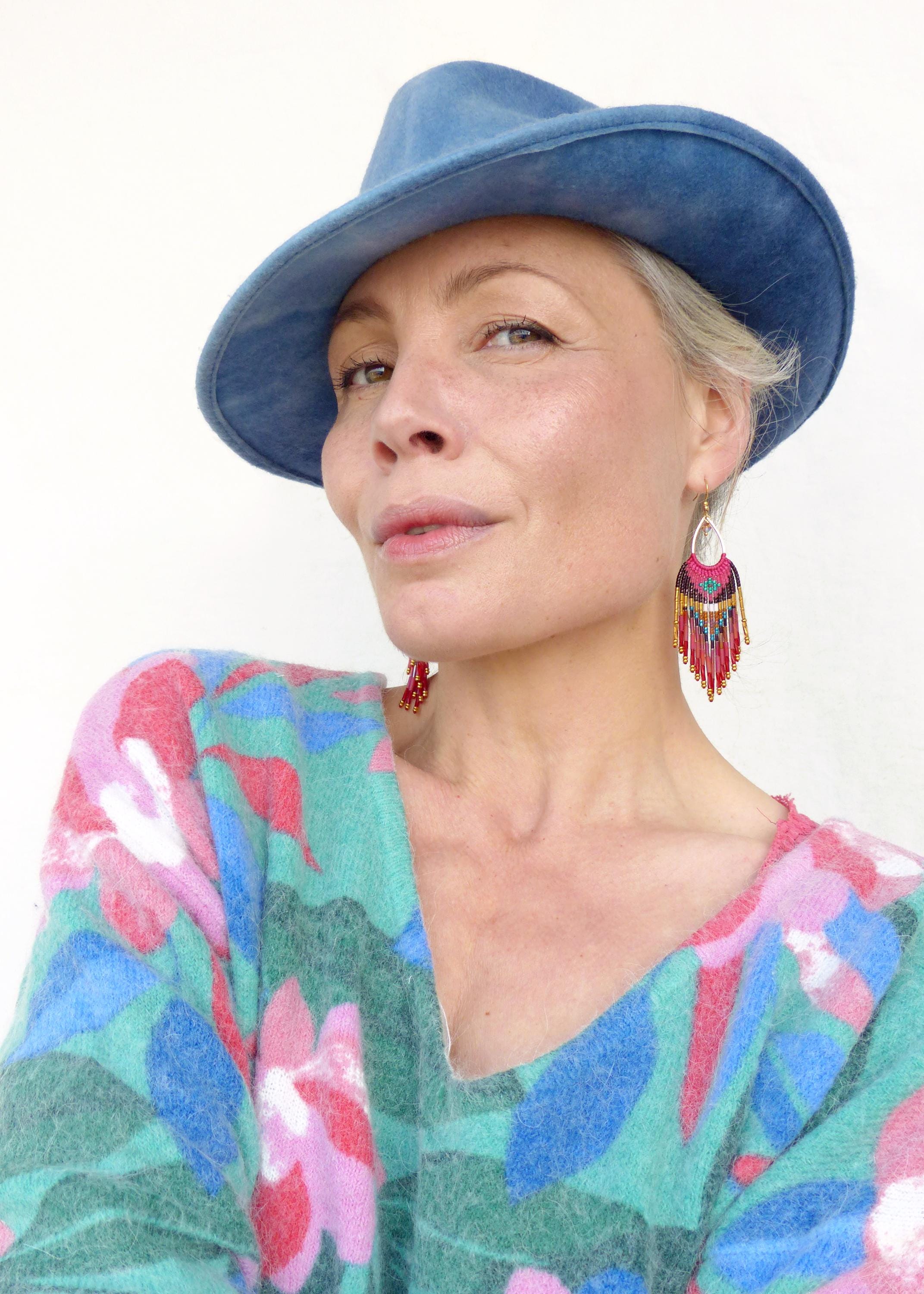Peut inclure: Une personne portant un chapeau bleu et des boucles d'oreilles colorées. Les boucles d'oreilles ont un dessus doré avec des franges rouges, roses et bleues. La personne porte également un pull avec un motif floral dans les tons de bleu, rose et vert.